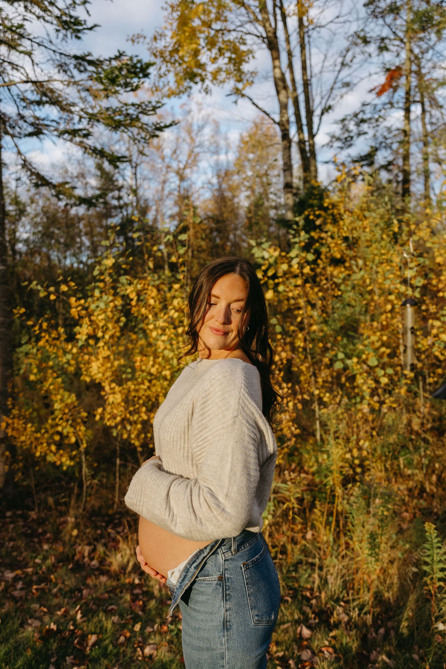 A person in a cream sweater and jeans poses outdoors against autumn foliage in golden sunlight.