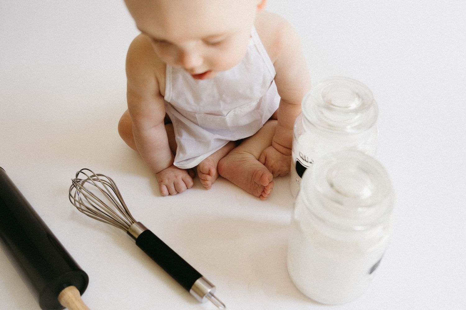 Baby in white diaper reaching for glass jar on white surface while adult hand holds fountain pen.