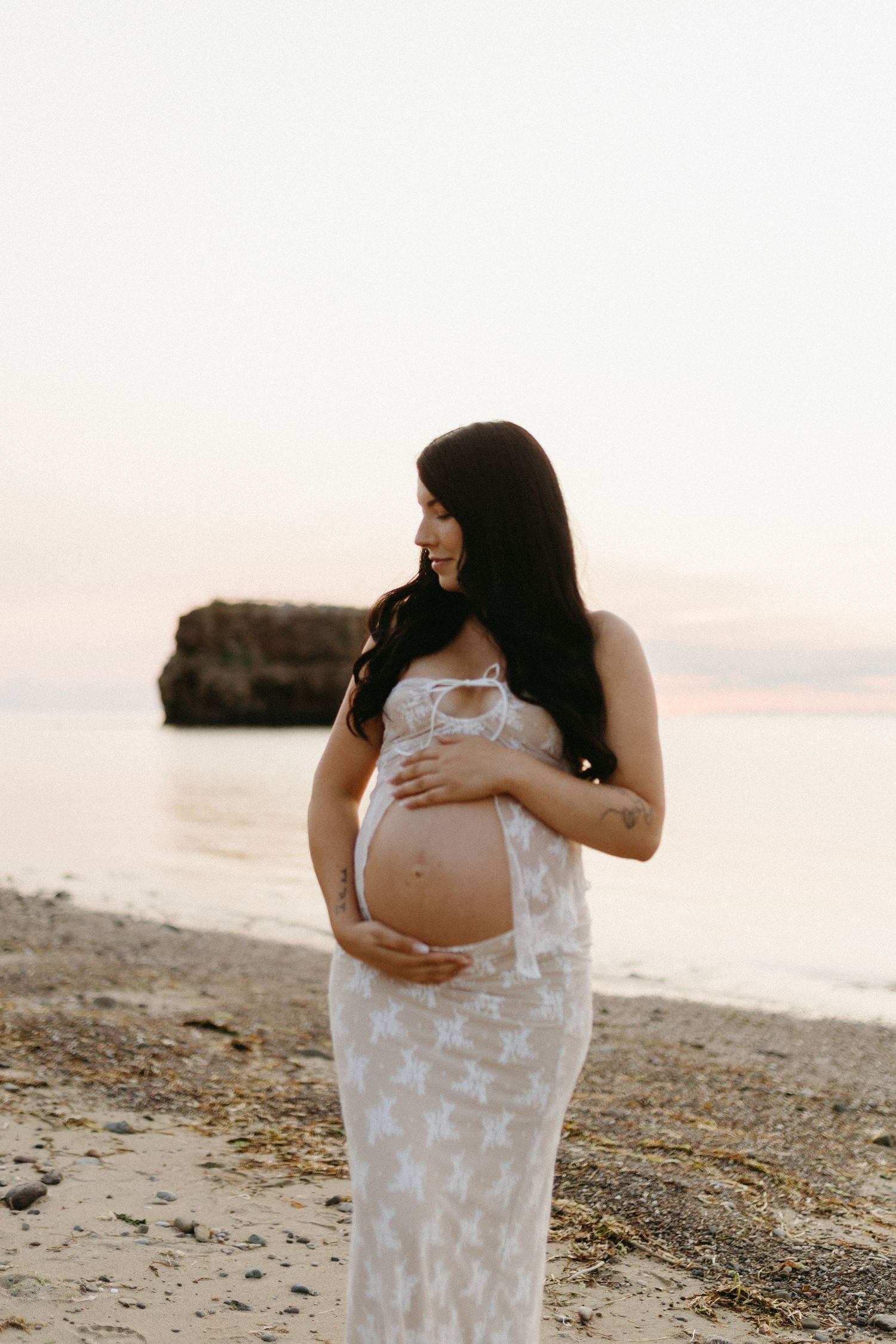 A pregnant woman in a white lace dress stands on a beach at sunset with ocean and rocks in the background.