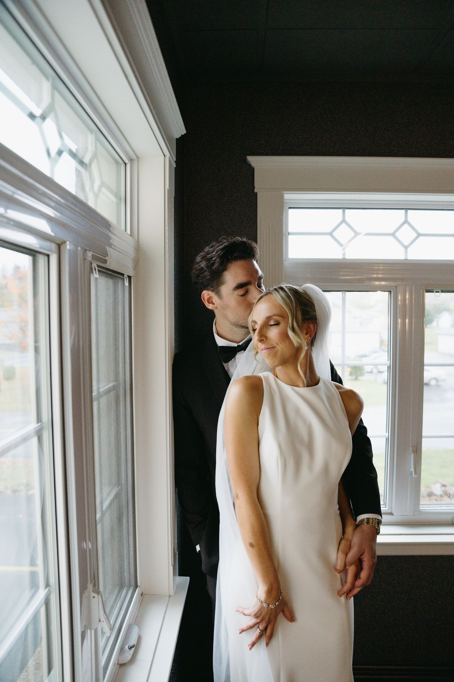 A minimalist wedding scene by large windows showing intimate moments between a couple in elegant attire.