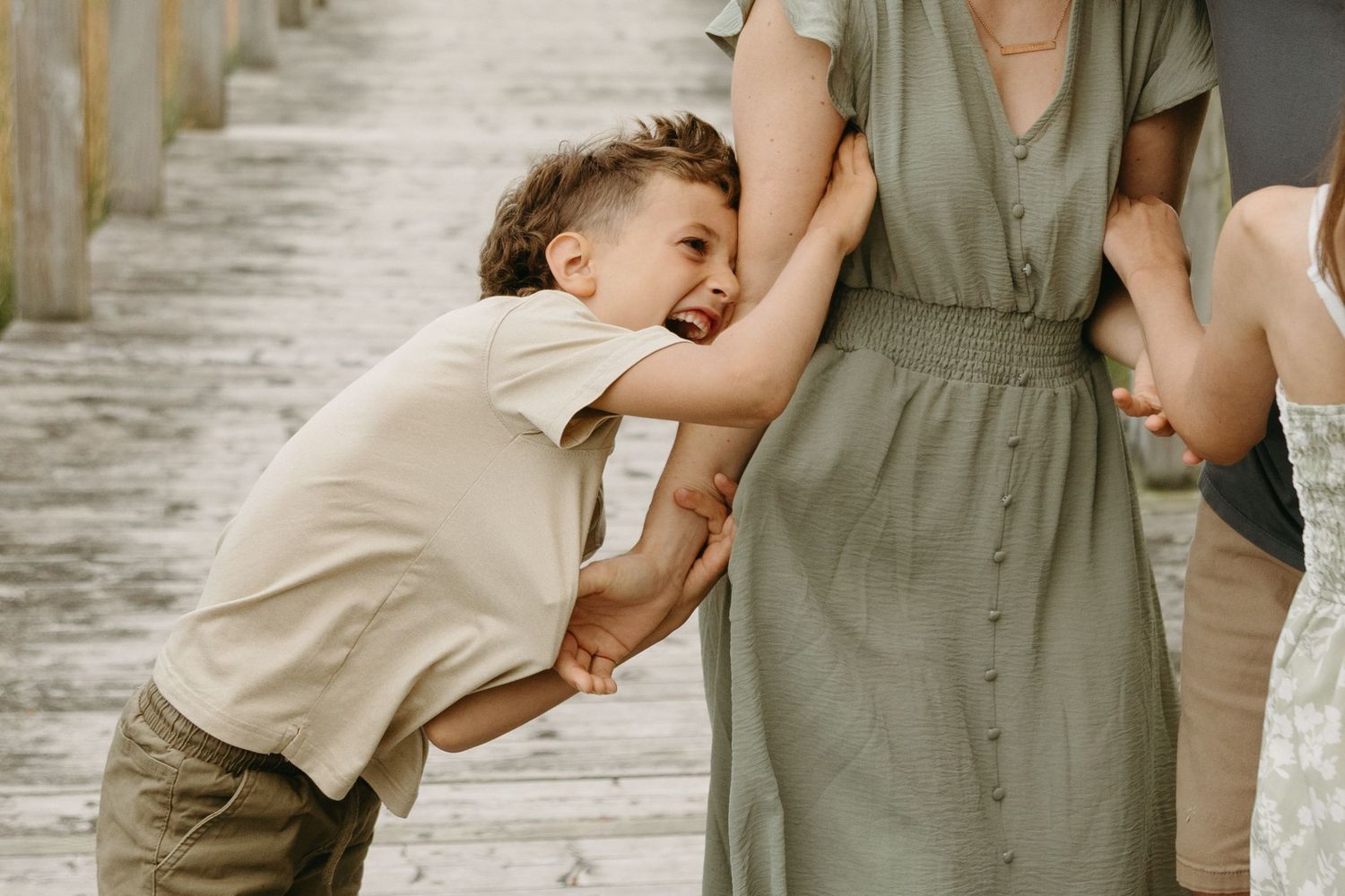 A child in beige clothing hugs someone wearing an olive green dress while standing on a concrete walkway.