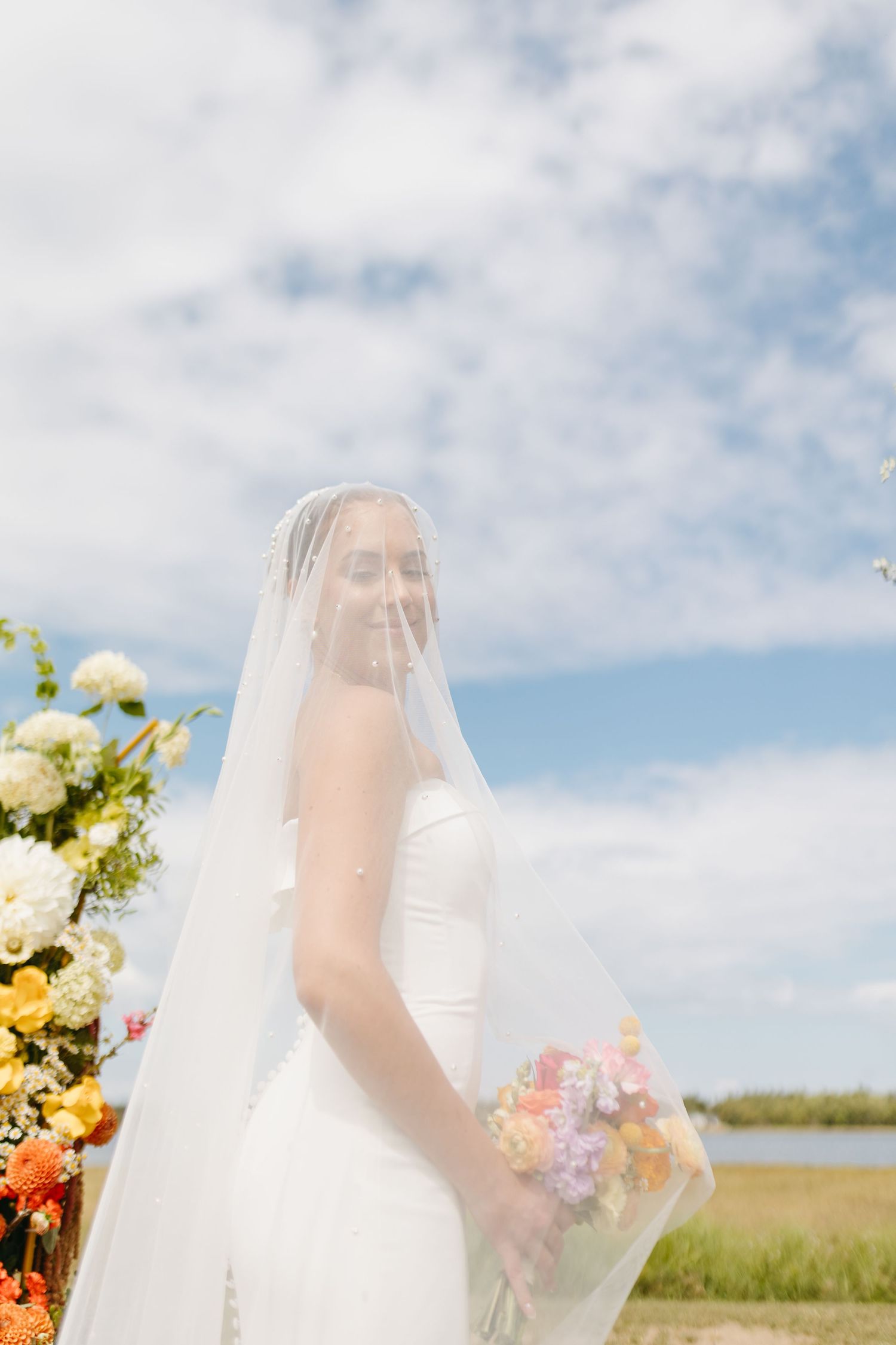 A bride in a white wedding dress and veil holds a bouquet of pink and white flowers against a blue sky.