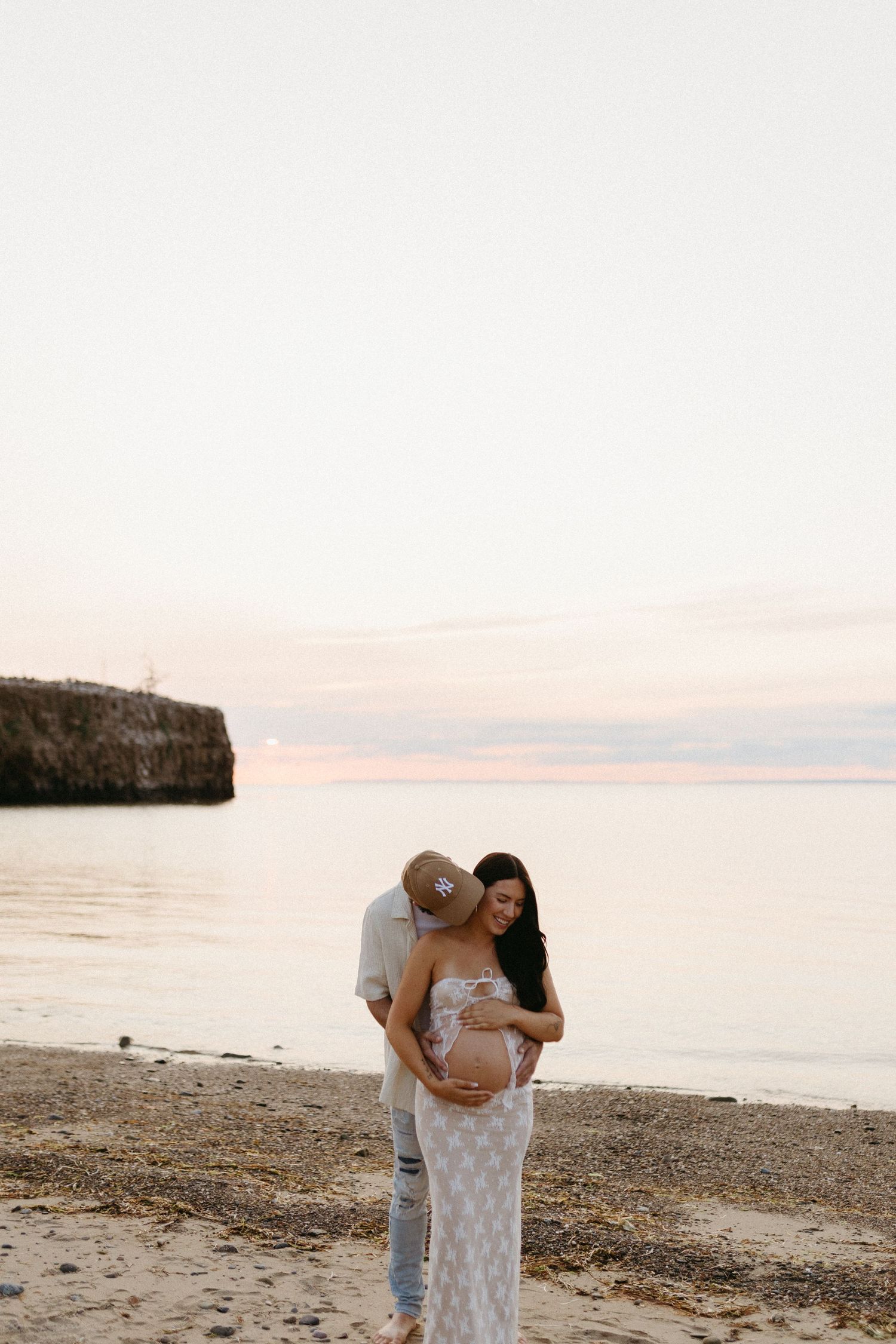 Couple embraces on peaceful beach at sunset with rocky cliff and calm ocean waters in background.