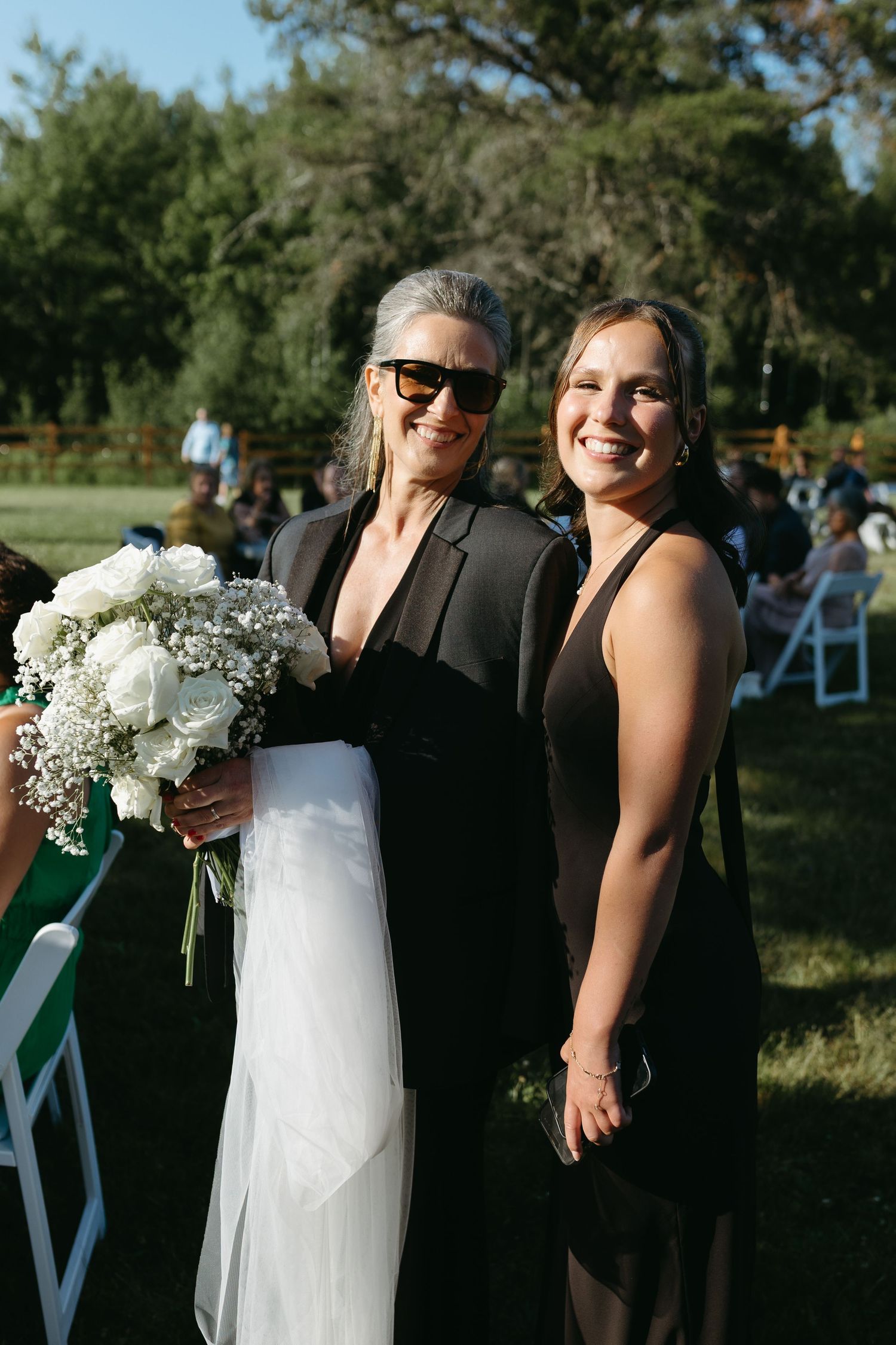 Wedding party members in black dresses share laughs and hugs while holding white floral bouquets outdoors.