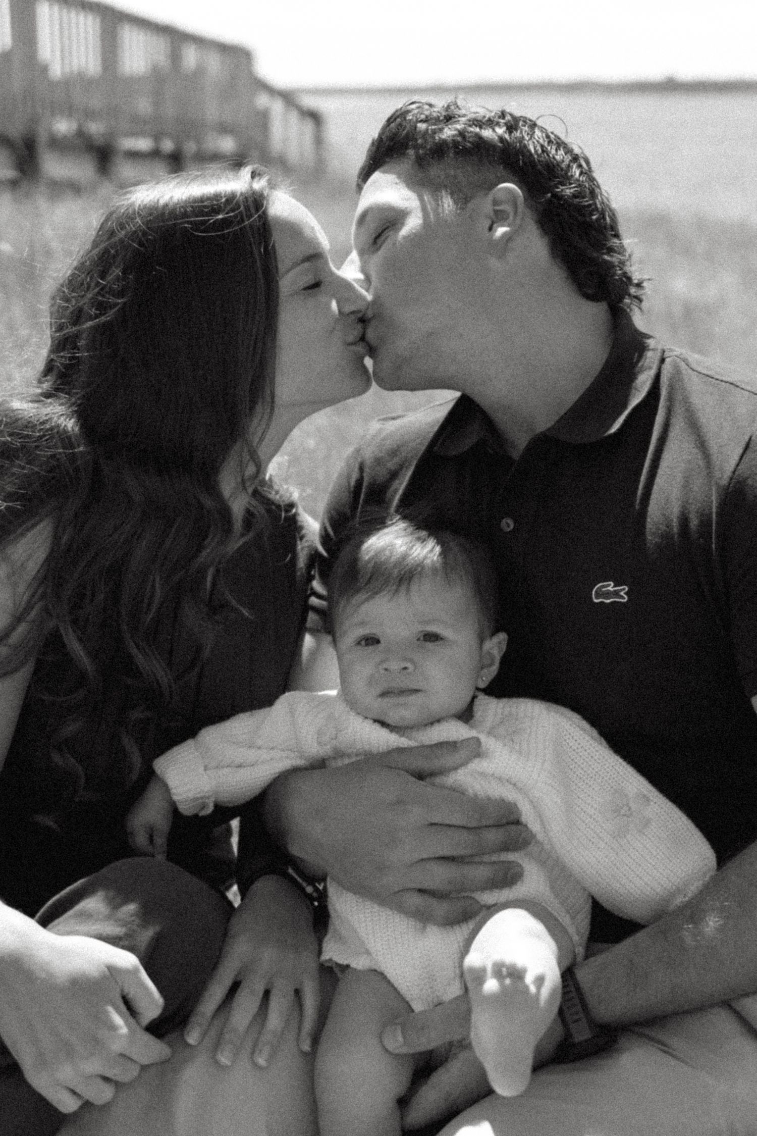A black and white intimate family portrait showing a couple sharing a kiss while holding their baby.