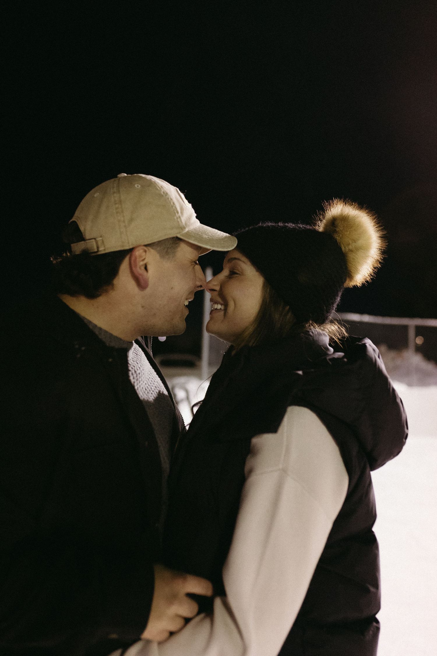 A couple sharing a romantic kiss in silhouette against a dark background while wearing winter coats and hats.