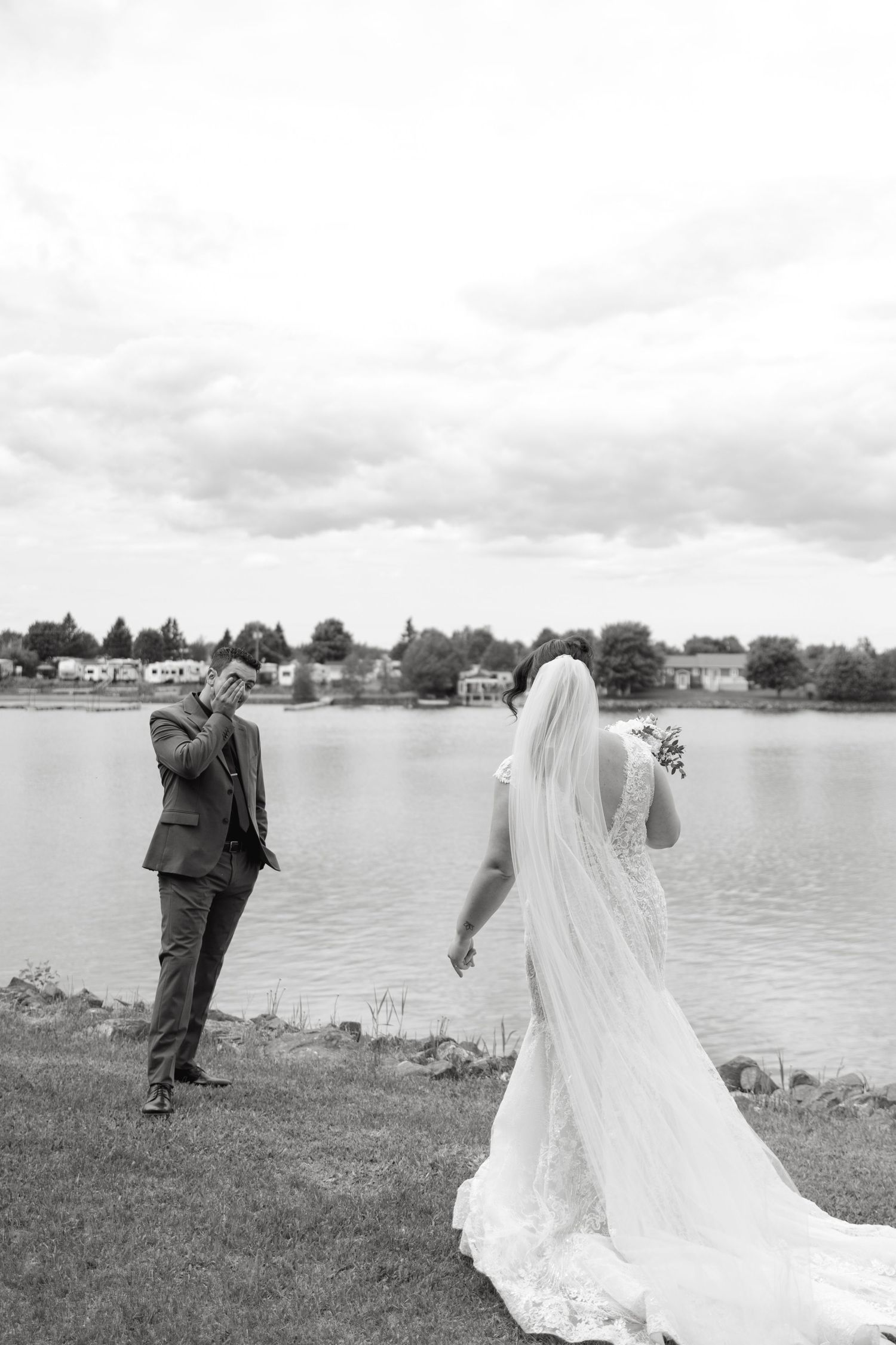 Black and white wedding photo showing a couple by a lake, with the bride in a flowing veil and groom in a suit.