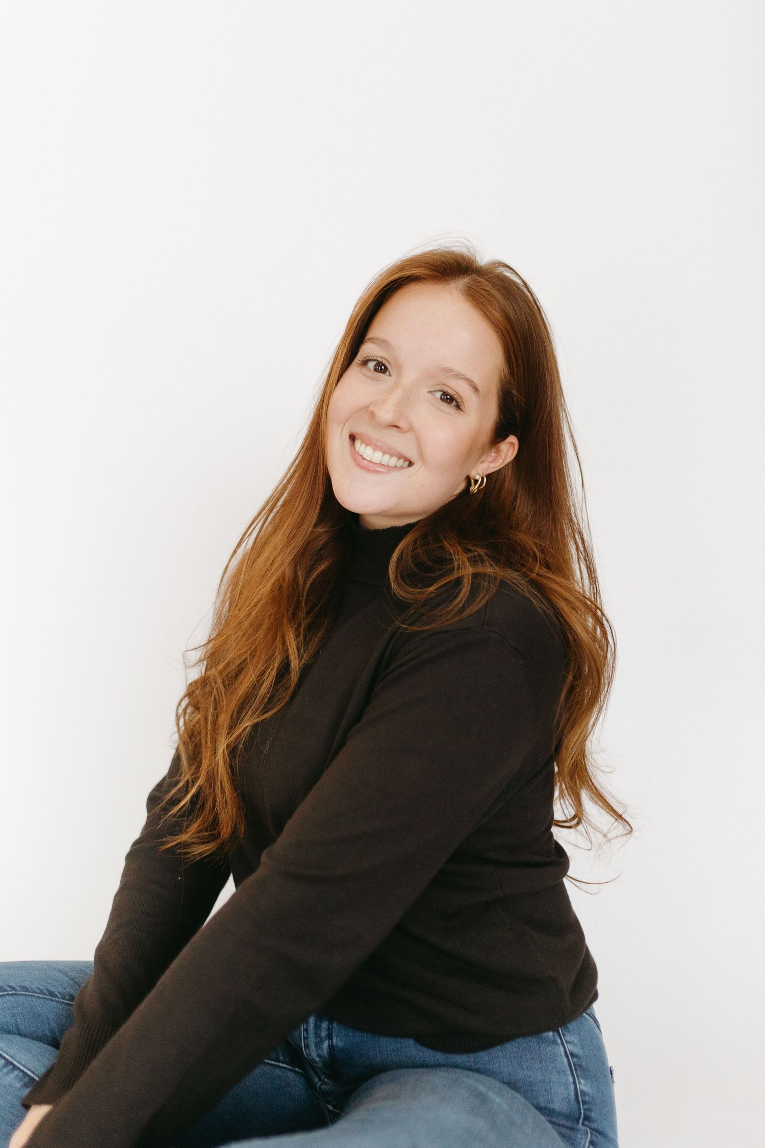 A person with red hair wearing a black turtleneck sweater and jeans poses against a white background.