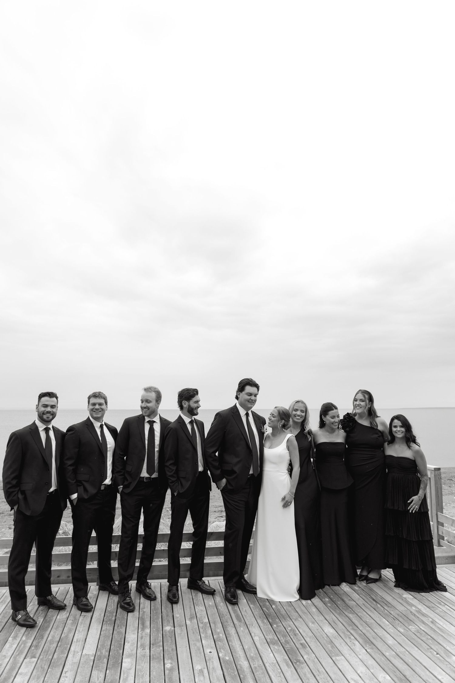 Wedding party in black and white attire poses together on a wooden deck overlooking the water.