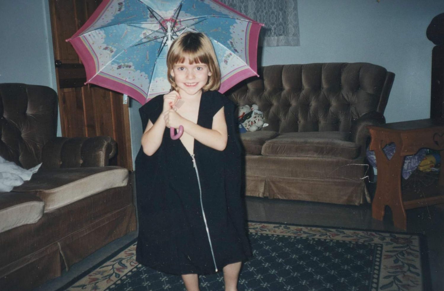 A person holds a pink umbrella while dancing and posing in a dimly lit living room with brown furniture.
