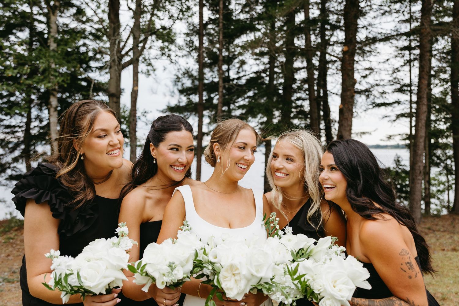 Bridesmaids in black dresses and white bouquets share joyful laughter together during outdoor wedding photos.