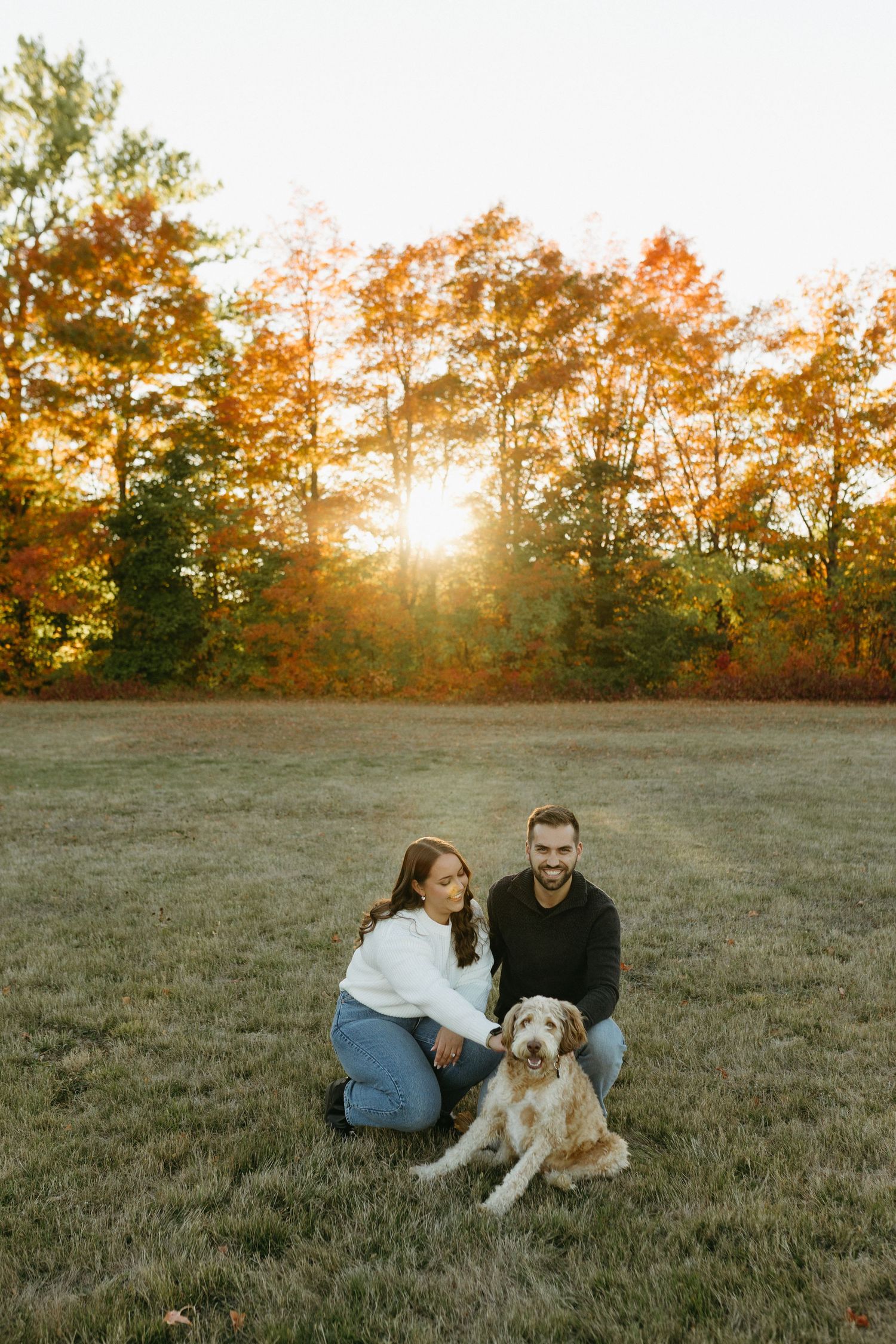 A couple sits with their golden retriever dog in a grassy field at sunset with autumn trees in the background.