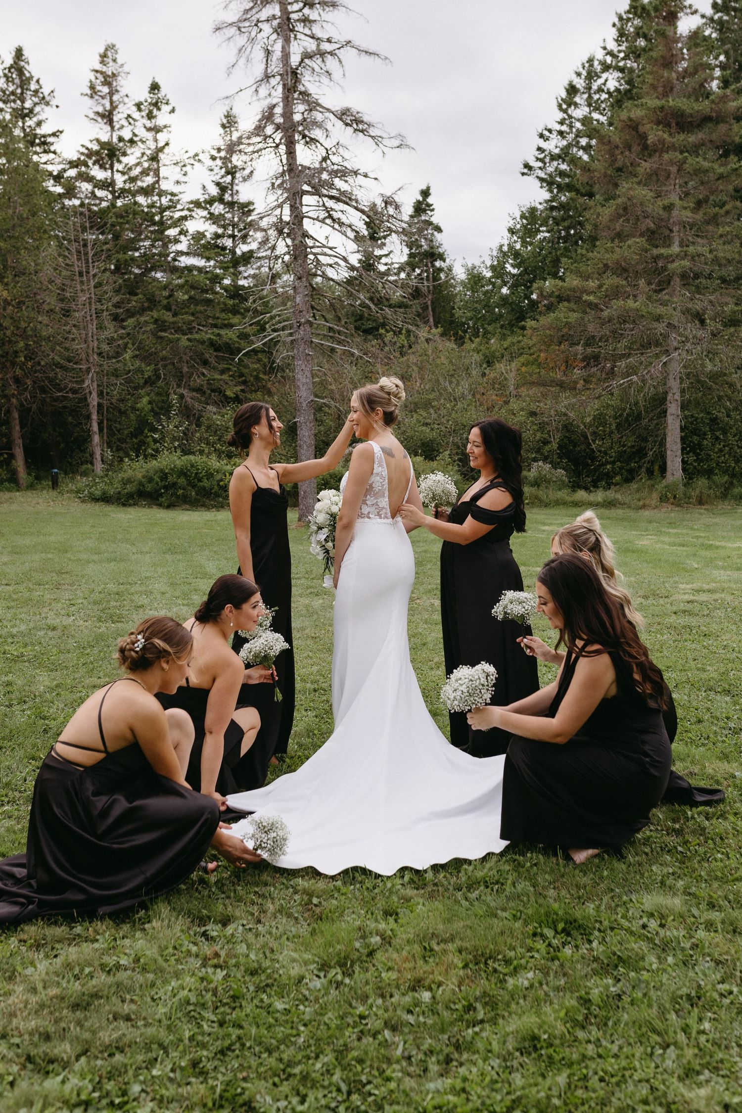 A wedding party in black dresses helps adjust a bride's flowing white gown on a grassy lawn surrounded by evergreen trees.