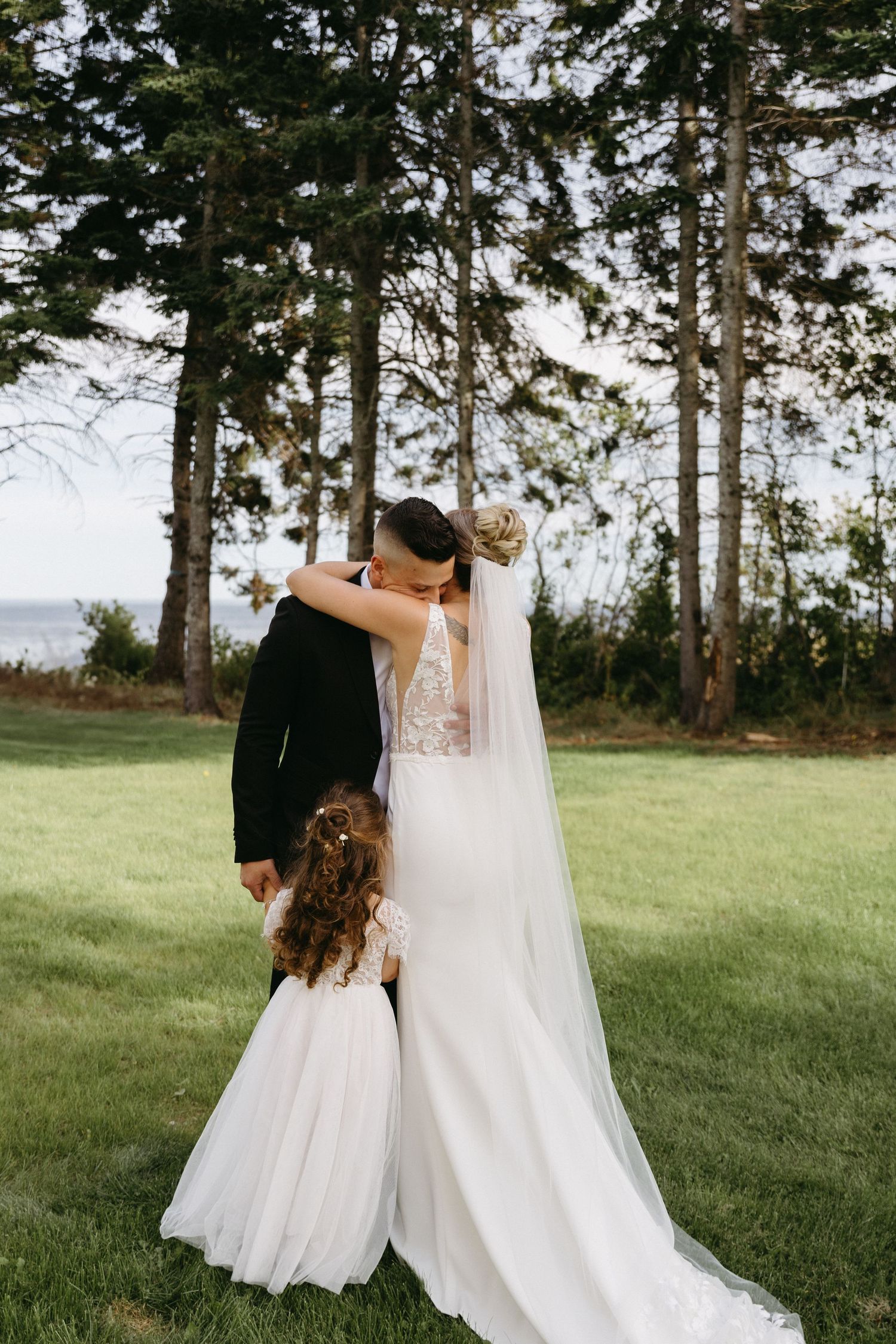 A touching moment between a bride in white and a young flower girl in a matching dress against a backdrop of tall pine trees.