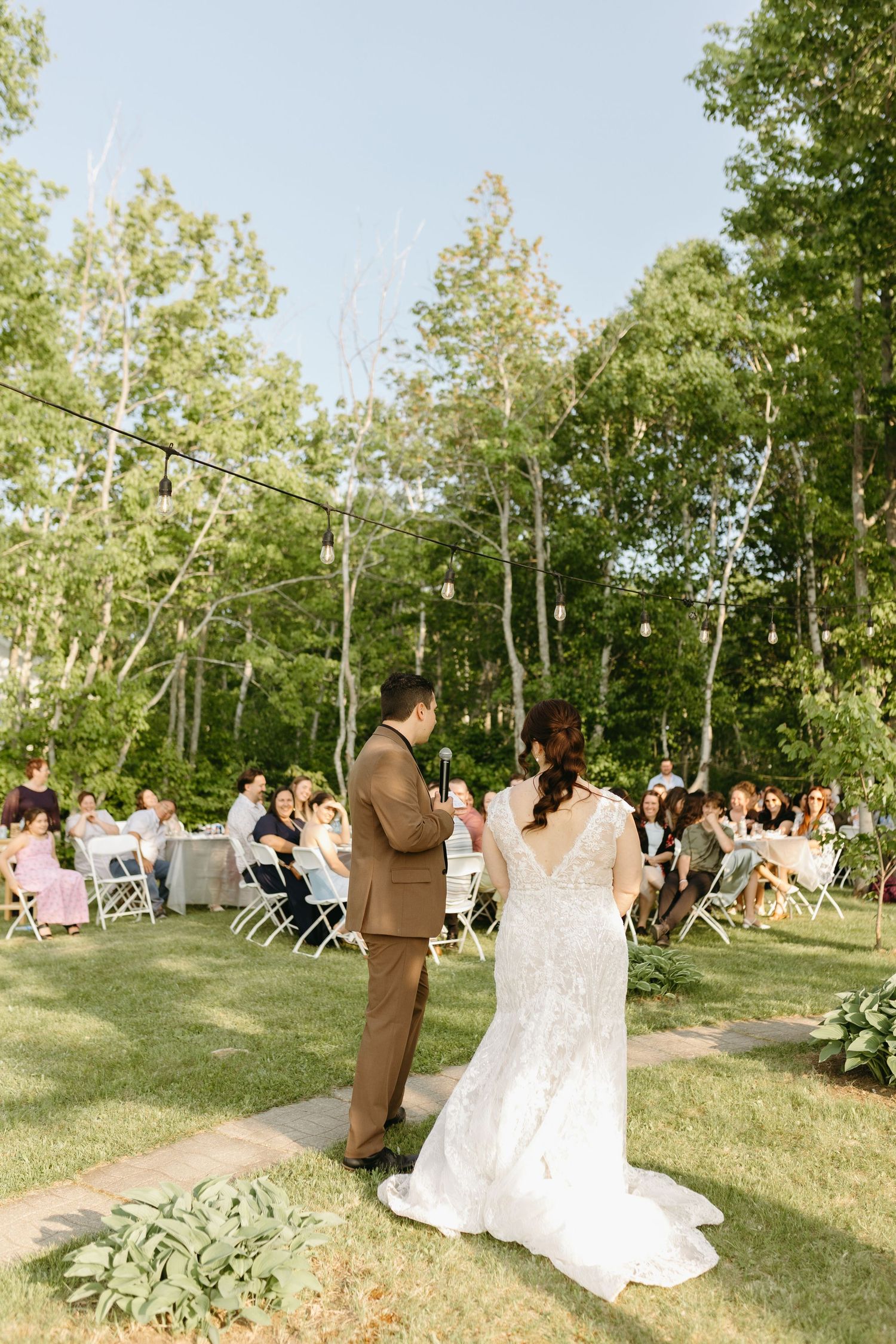 A couple stands together during an outdoor wedding ceremony in a lush garden setting with guests seated on white chairs.