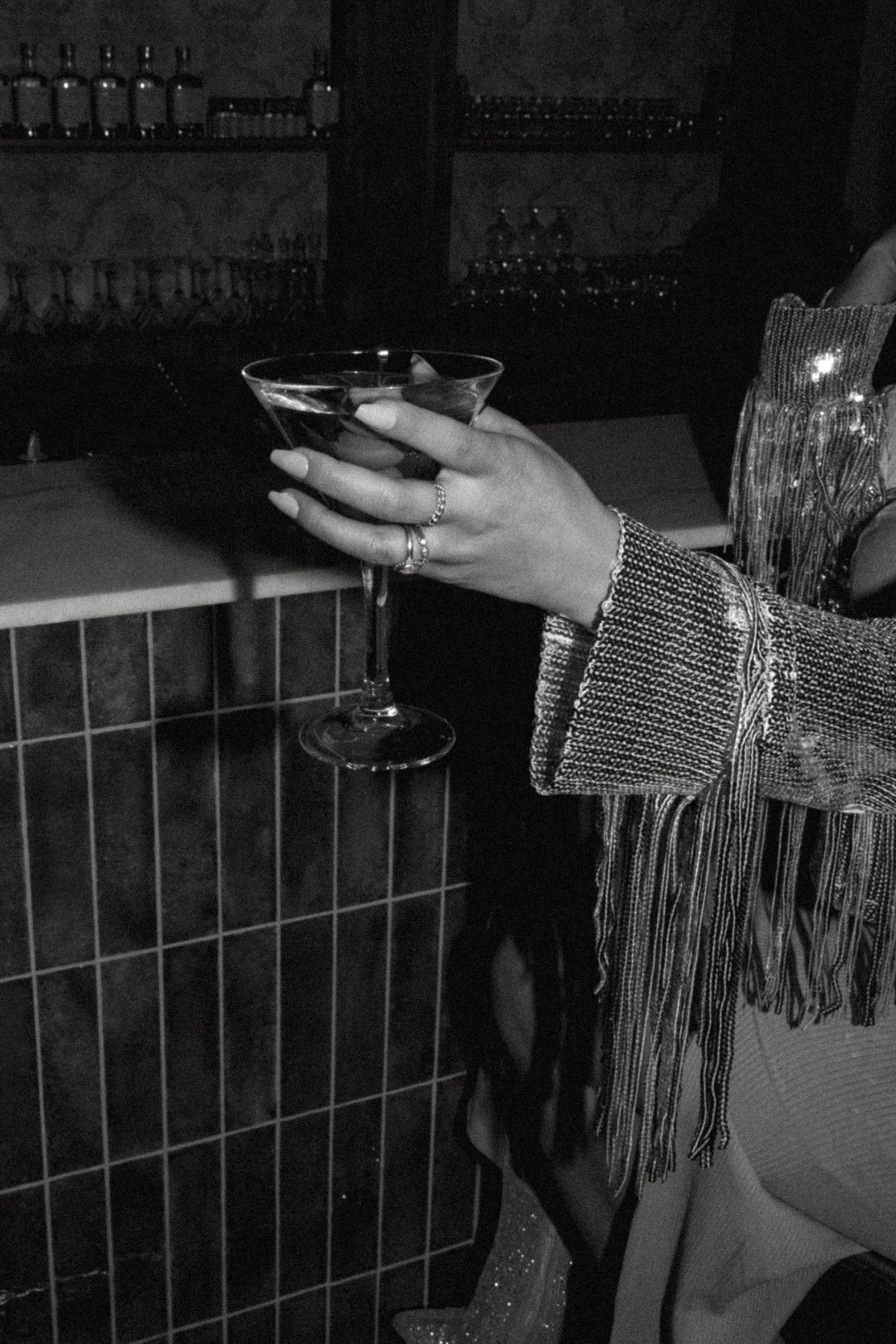 Hand holding martini glass with fringe sleeve detail in dark bar setting.