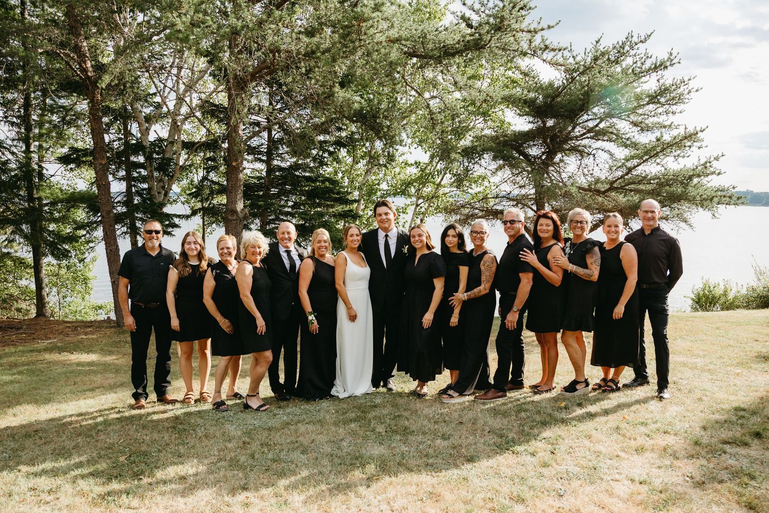 Wedding party members in black and white attire pose together outdoors on a grassy hillside during a sunny day.