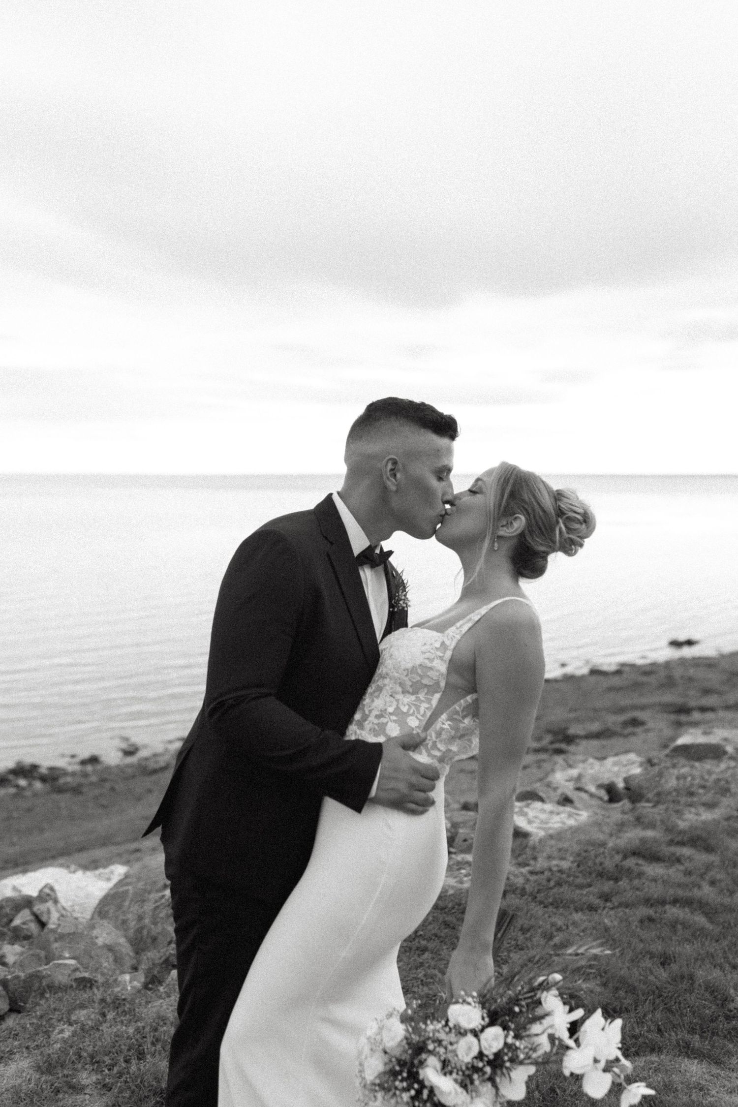 A romantic black and white wedding photo of a couple kissing on a coastal cliff overlooking the ocean.
