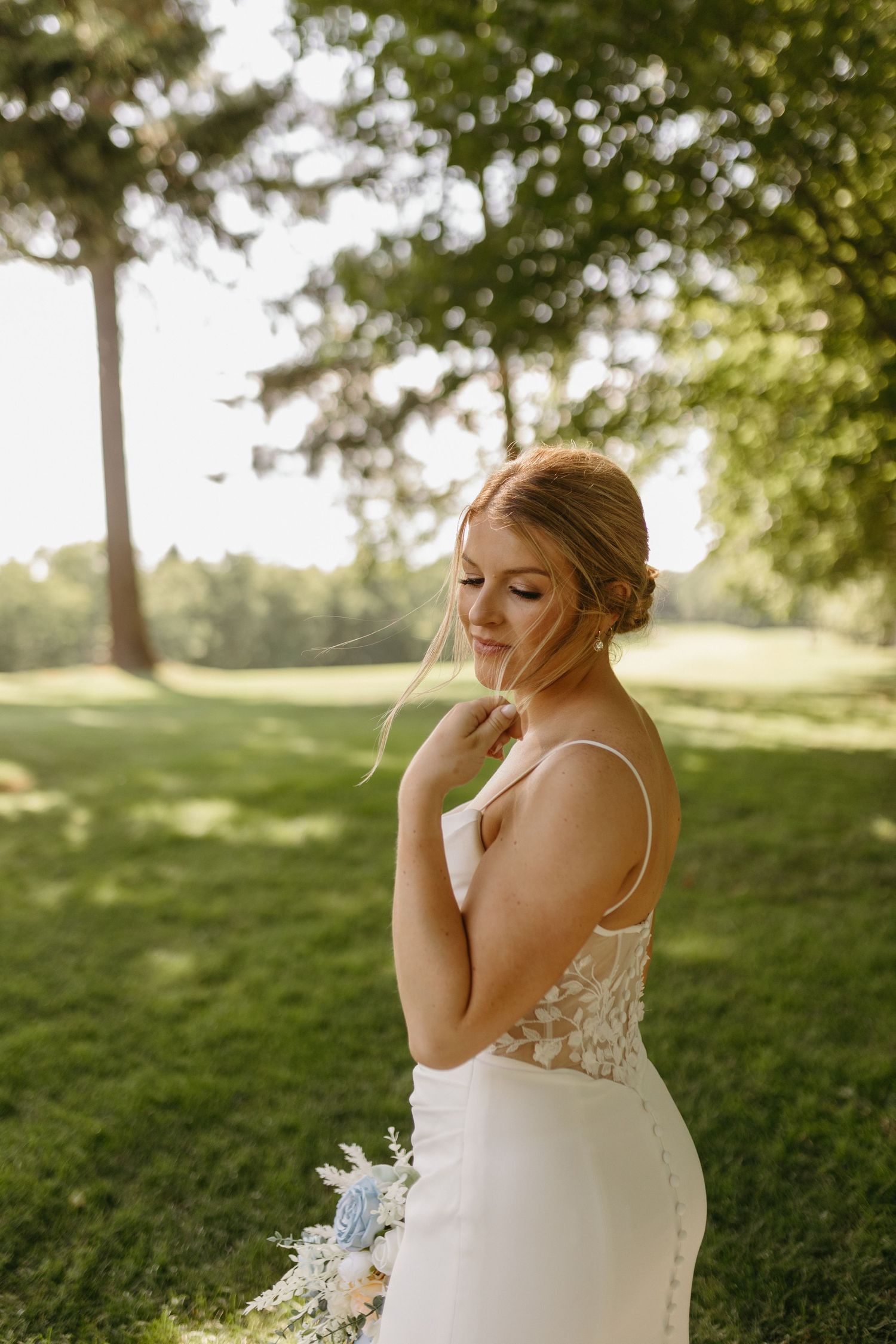 Bride in lace dress stands on grassy hill during golden hour outdoor wedding photoshoot.