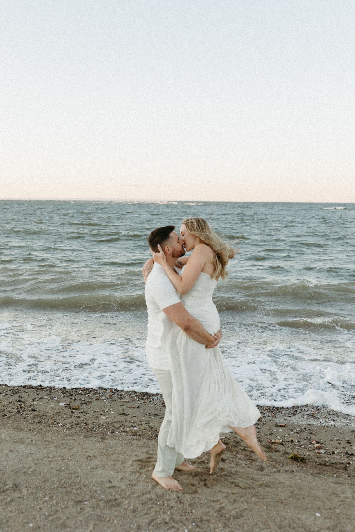 A couple in white clothing embraces on a beach at sunset.