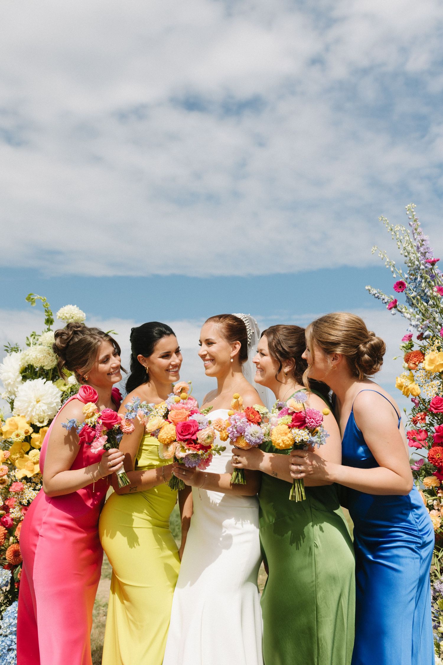 Bridesmaids in colorful dresses laugh together while holding vibrant bouquets against a blue summer sky.