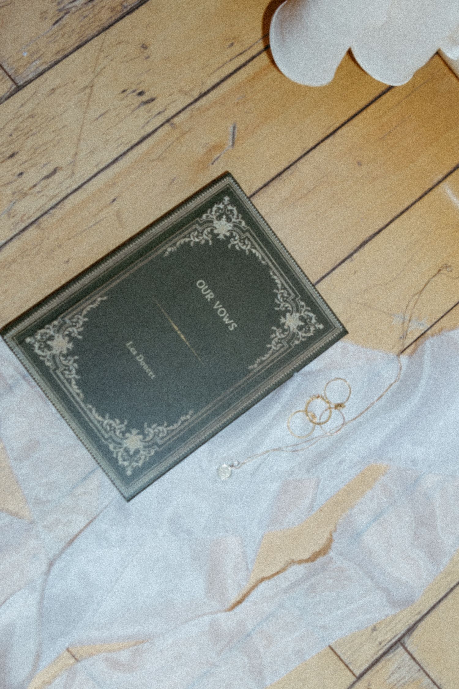 A vintage dark green book with ornate decorative details lies on a white cloth against wooden floorboards.