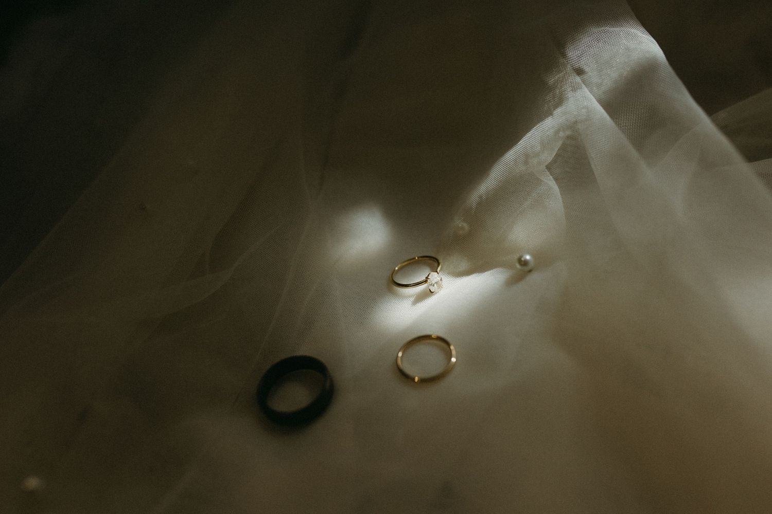 Wedding rings and veil photographed in dramatic moody lighting with soft focus and ethereal atmosphere.