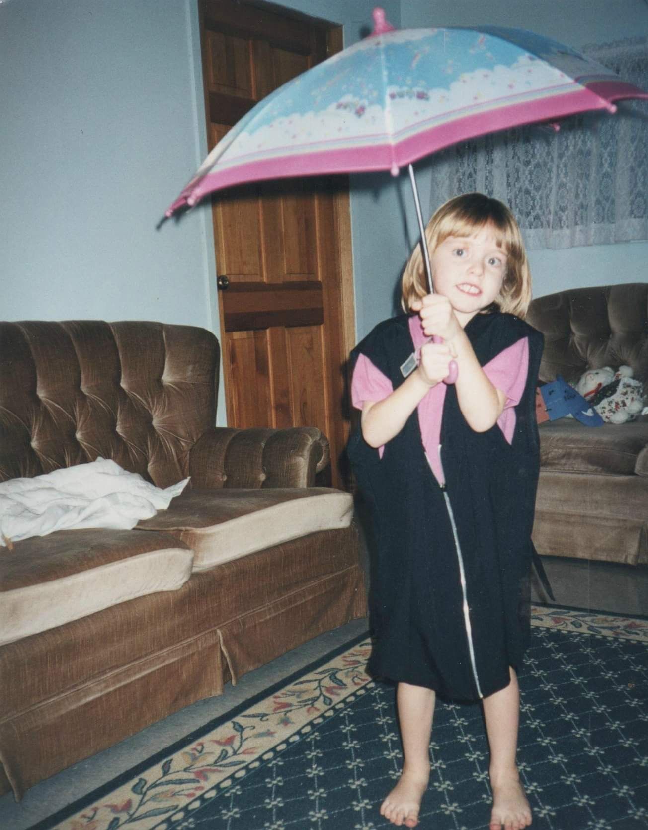A child in a black dress holds a pink and white umbrella while standing barefoot on a patterned rug in a living room.