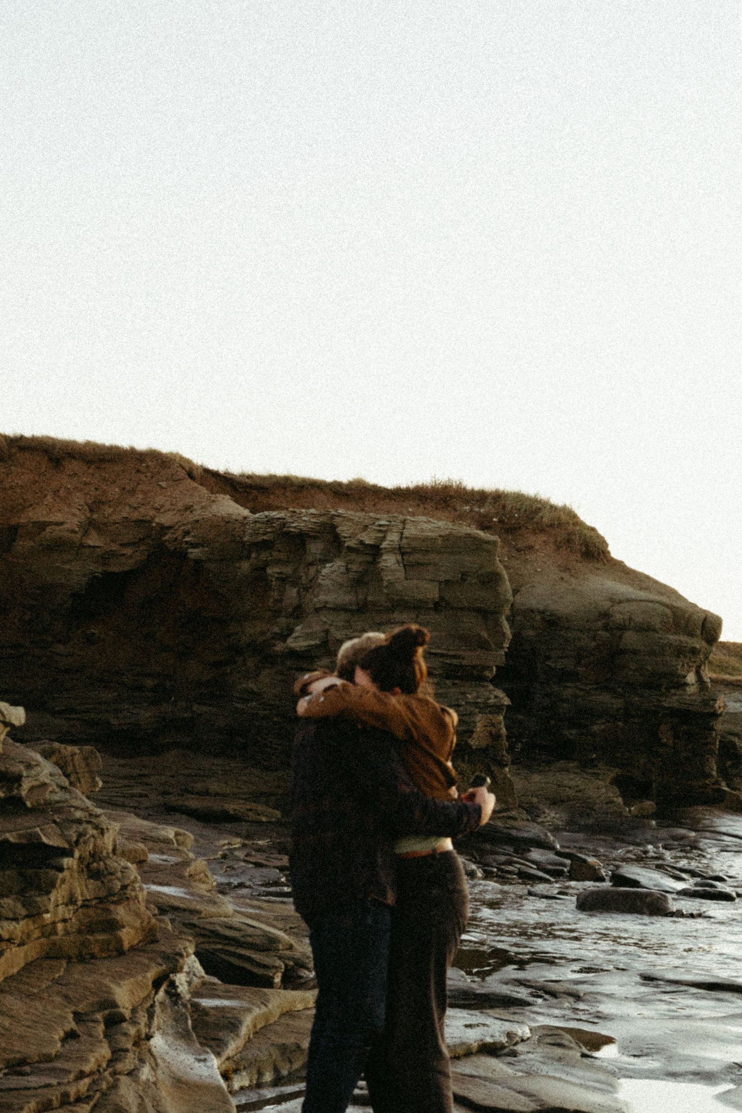 Two people embrace while standing on rocky coastal cliffs at sunset near the ocean.