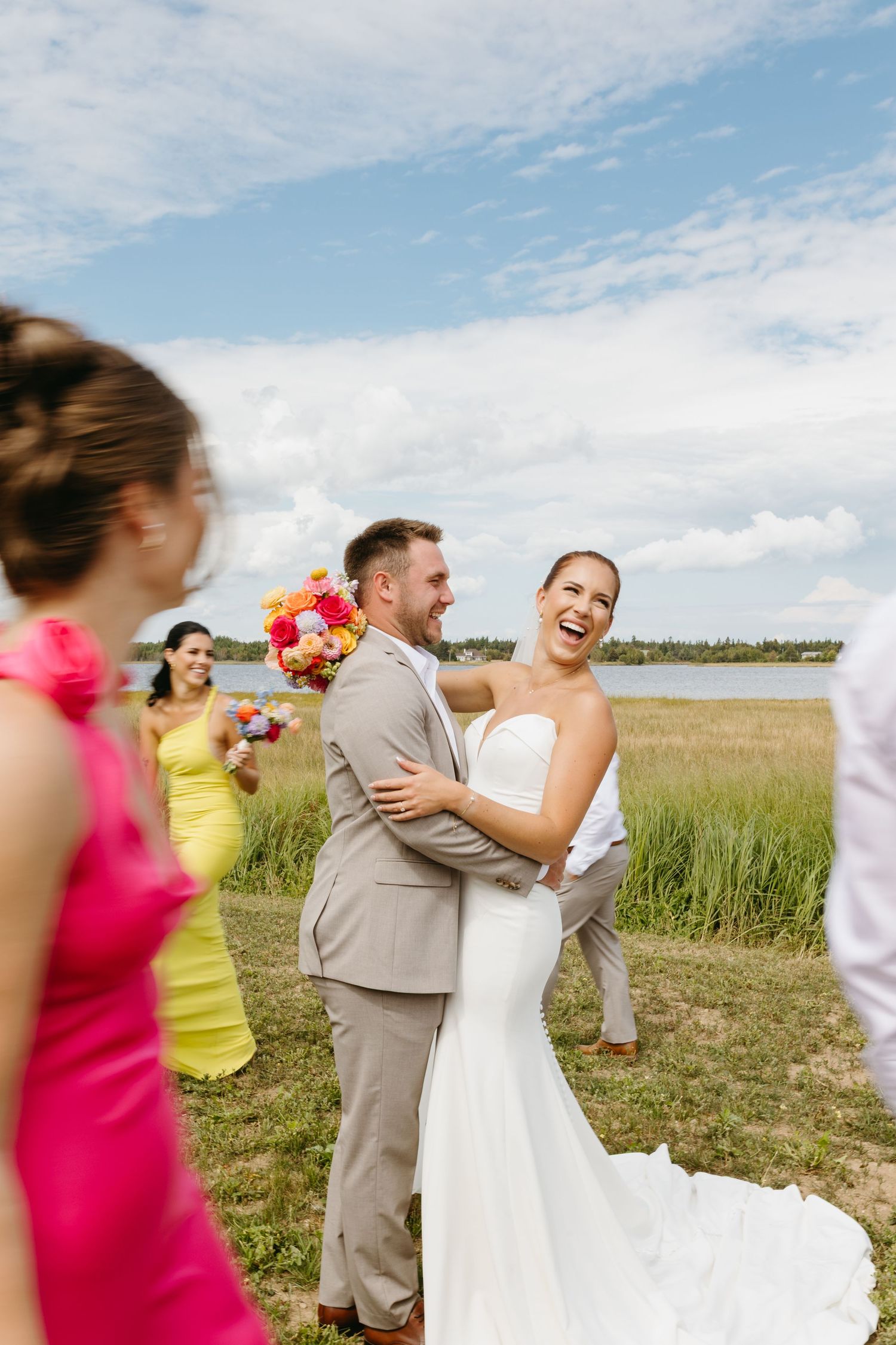 Wedding party celebrates joyfully in a sunny field with bridesmaids in bright pink dresses and groomsmen in gray suits.