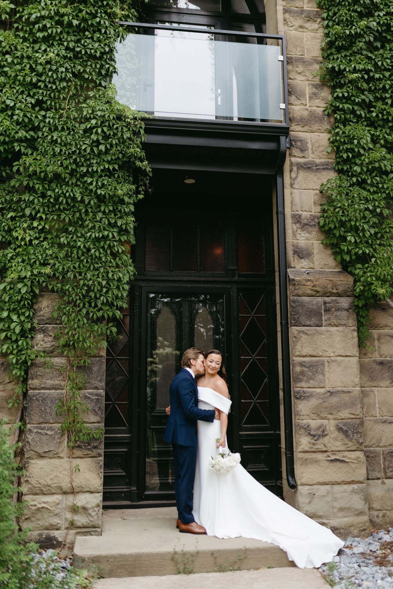 A couple in wedding attire share an intimate moment in front of an ivy-covered stone building entrance.