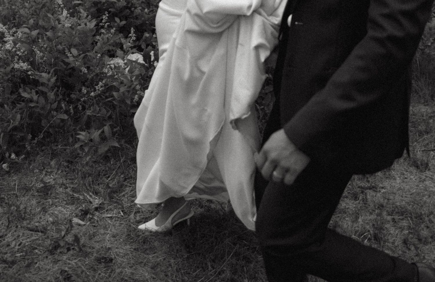 A black and white photograph showing the bottom portion of a wedding dress and suit at an outdoor ceremony.