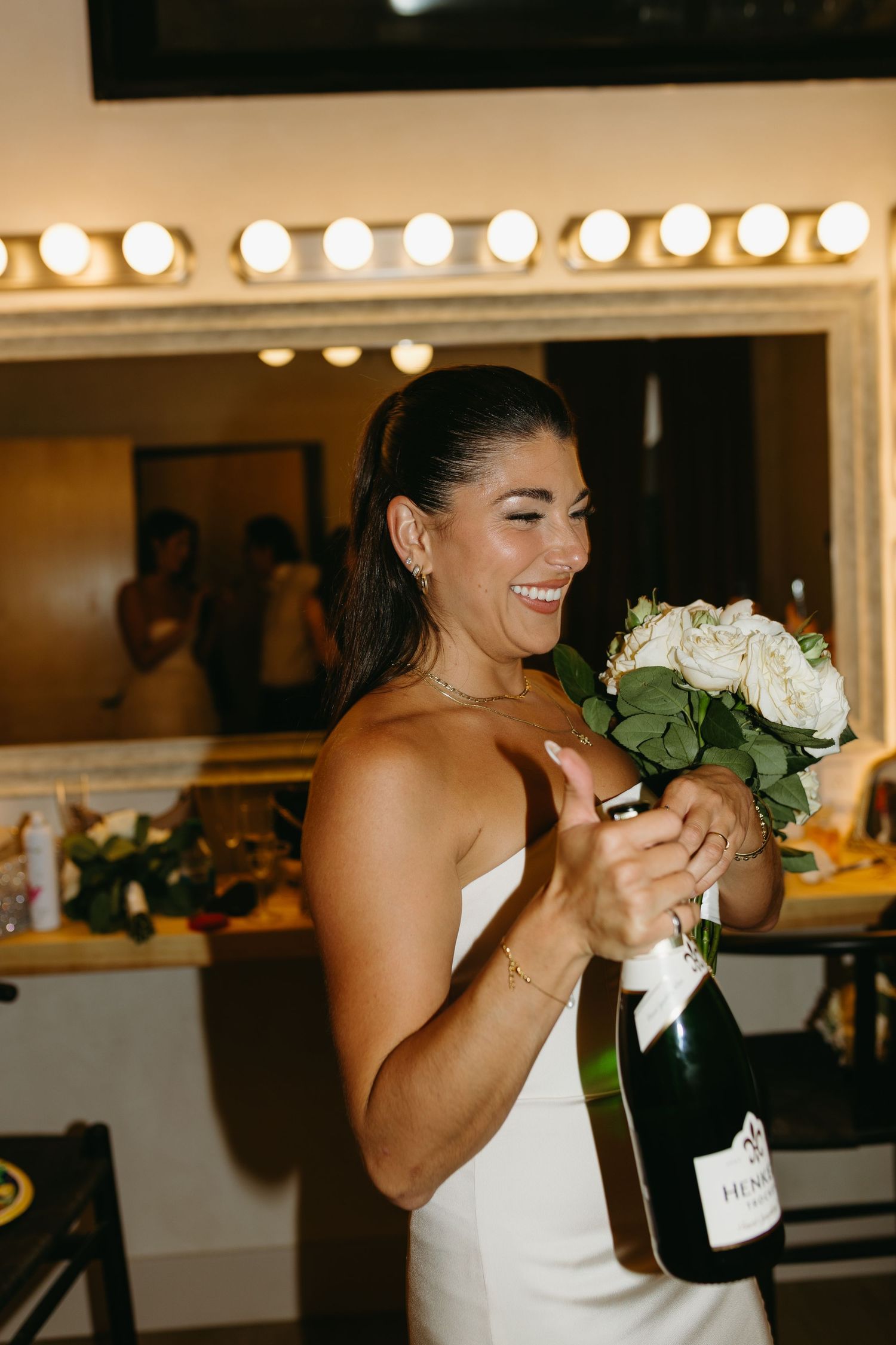 A joyful moment in a dressing room with champagne and white roses, illuminated by vanity lights.
