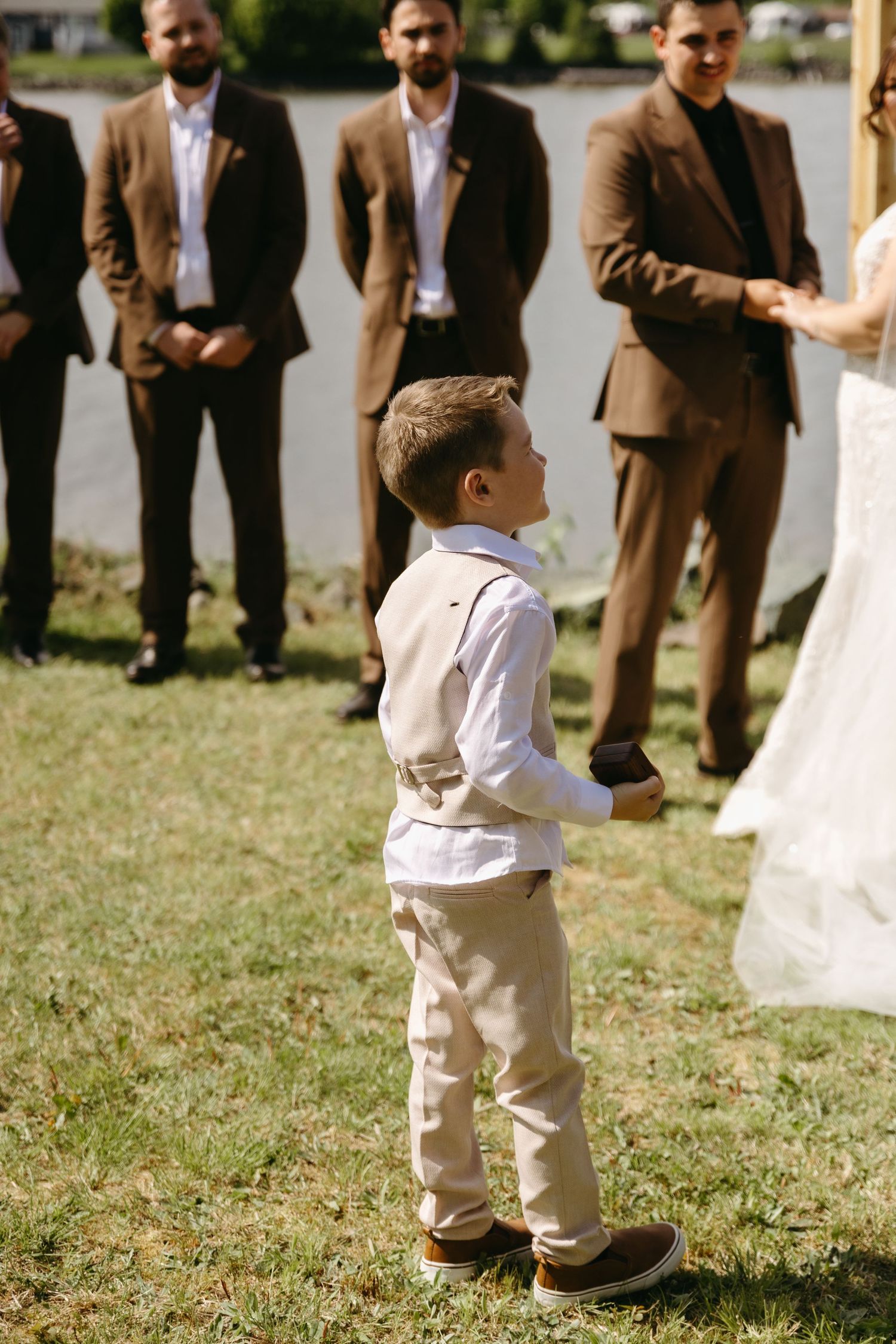 A young ring bearer in a white shirt and khaki pants stands at attention during an outdoor wedding ceremony.