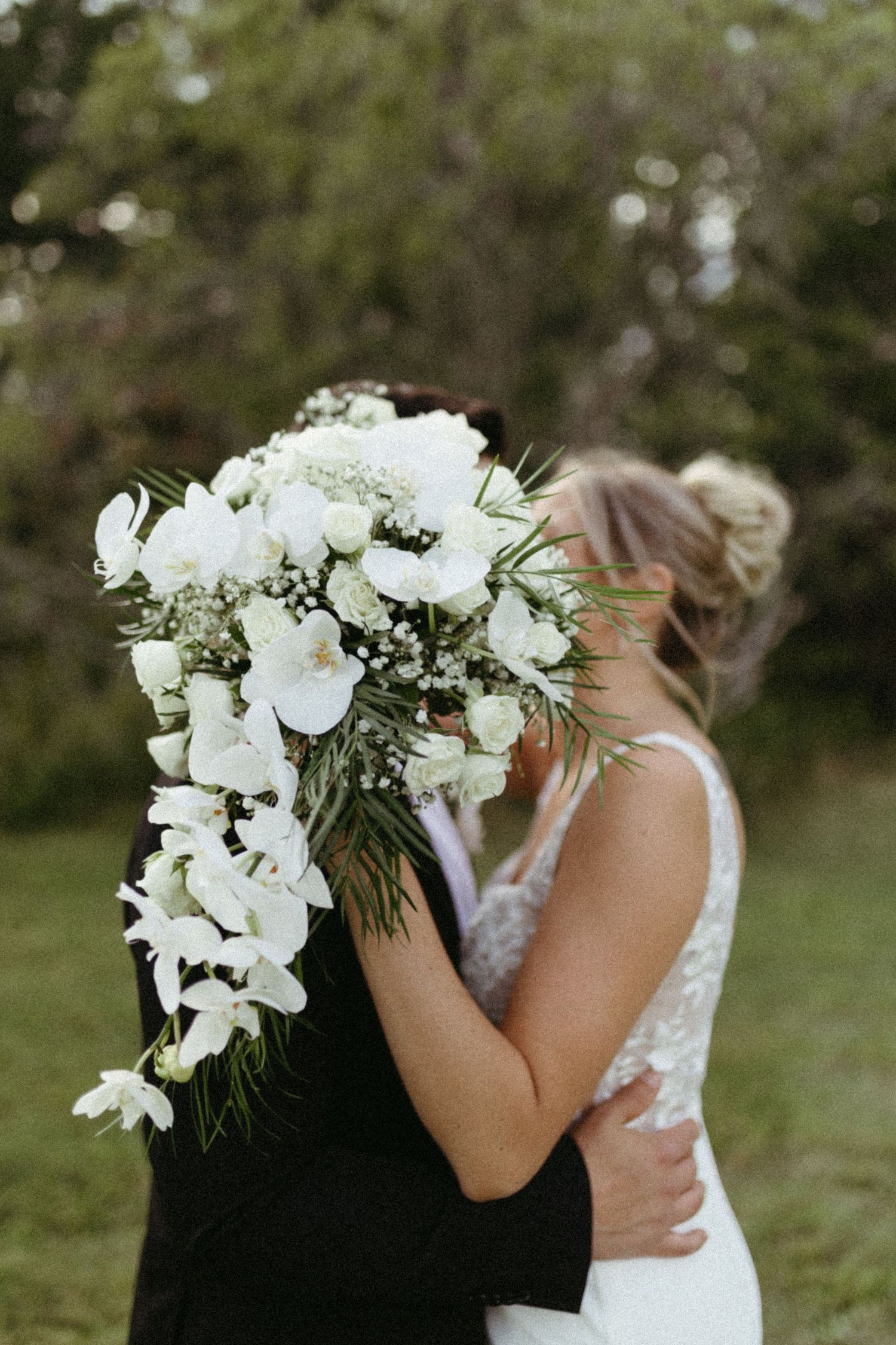 Two people share an intimate embrace while one holds a cascading white orchid bouquet in an outdoor garden setting.
