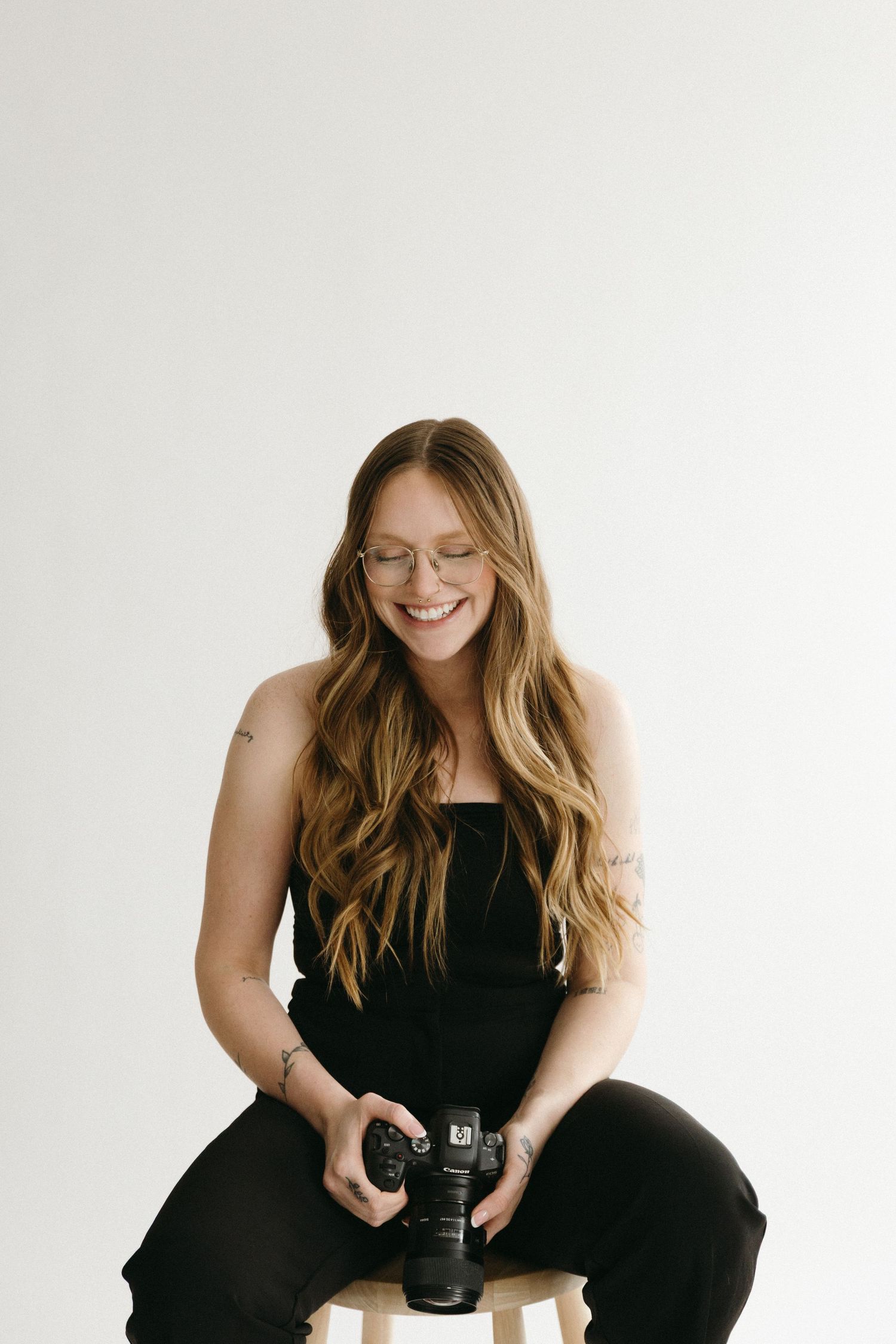 Professional photographer in black outfit sits on white backdrop while holding camera and smiling during photo session.