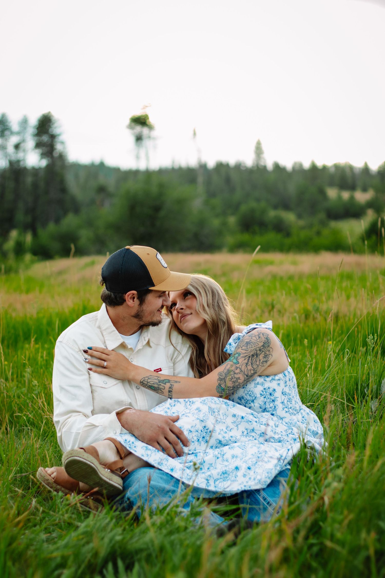 Couple sitting in lush grass sharing a tender moment, woman in blue floral dress with tattoos.