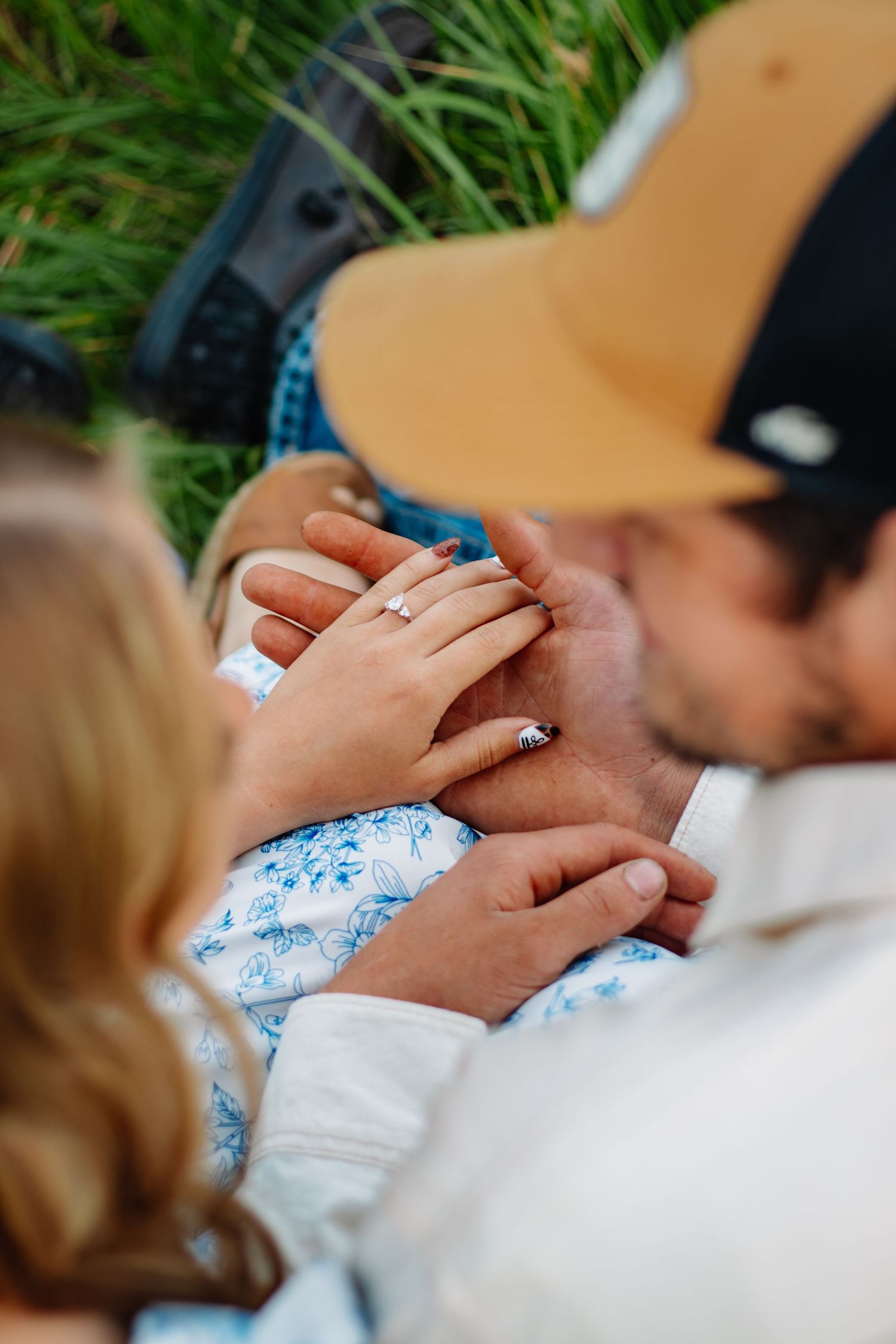 Close-up of couples intertwined hands with engagement ring while sitting in green grass together.