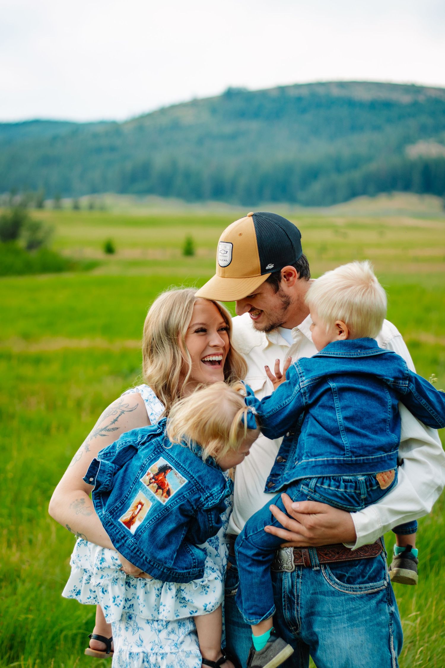 Laughing family with two young children in denim outfits enjoying green mountain field.