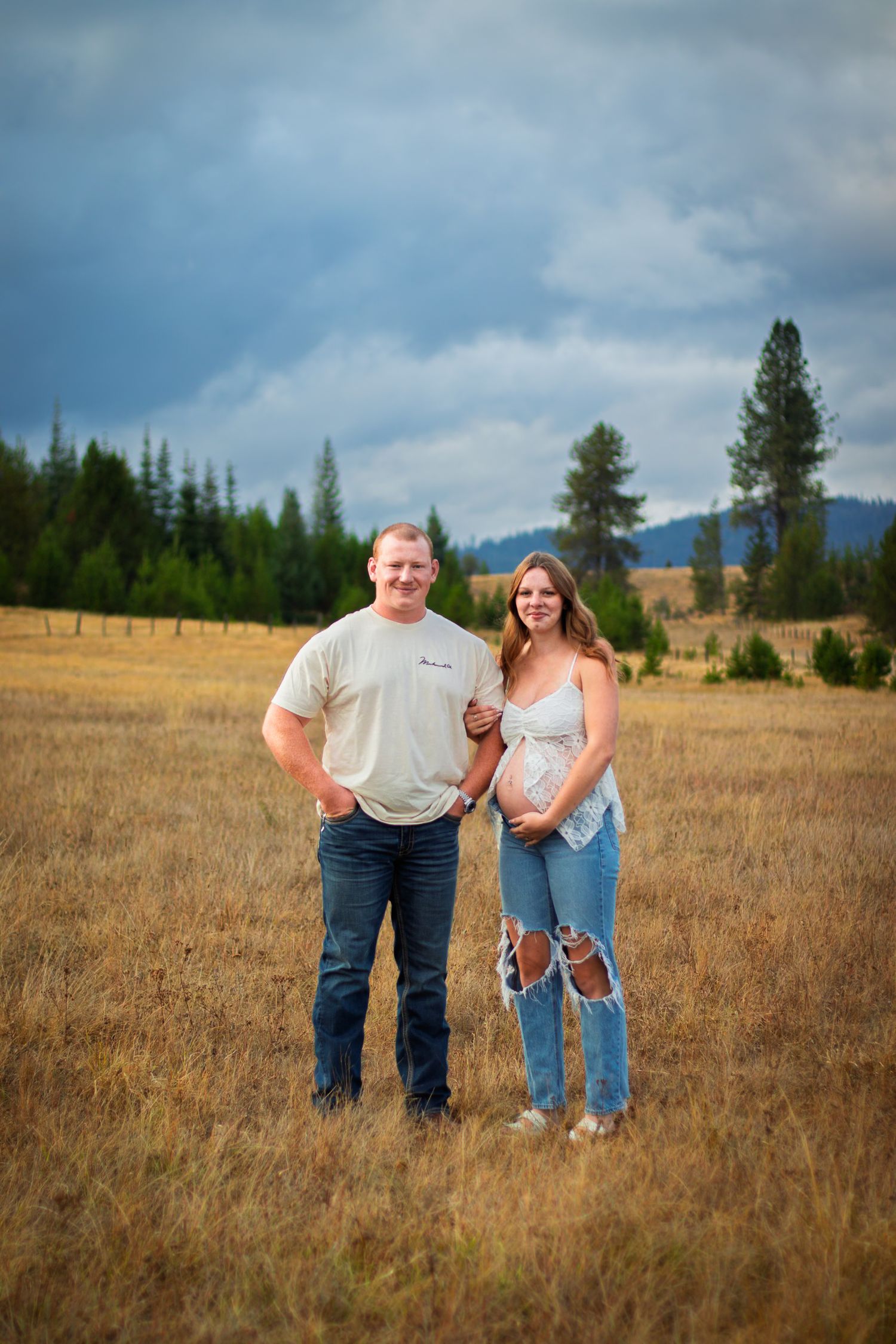Expecting couple posing together in a golden field under dramatic cloudy skies, woman cradling baby bump.