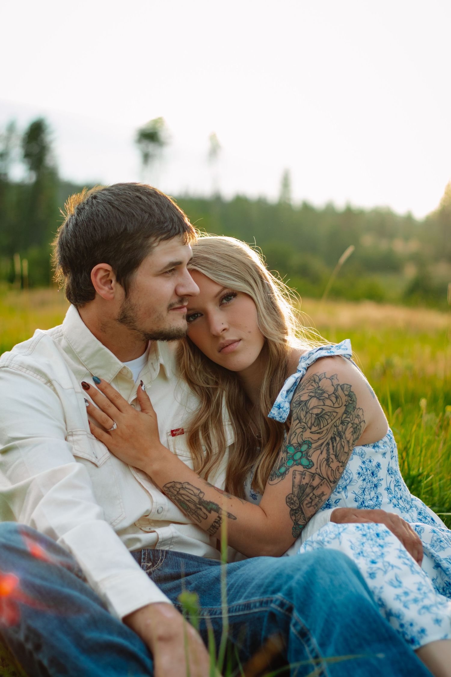 Couple sitting in a green meadow at sunset, woman with tattoos leaning on mans shoulder.