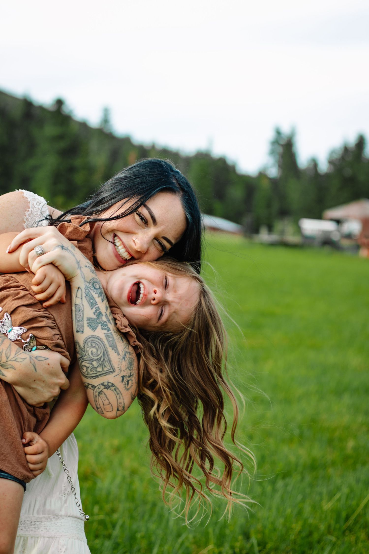Tattooed mother joyfully embracing her laughing daughter in a lush green field on a cloudy day.