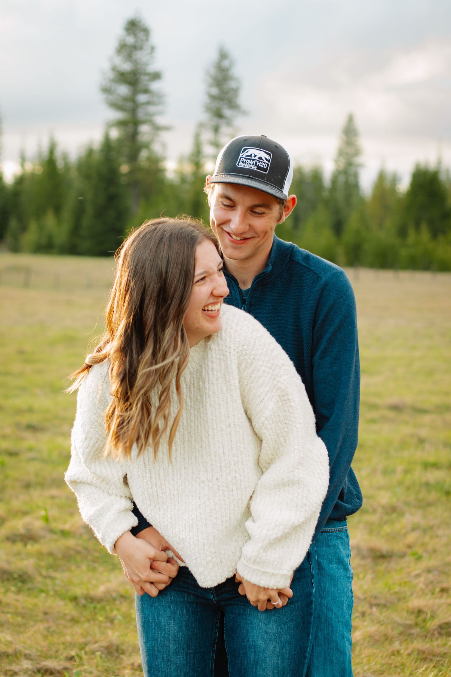 Happy couple embracing in green meadow, woman in white sweater laughing with pine trees behind them.