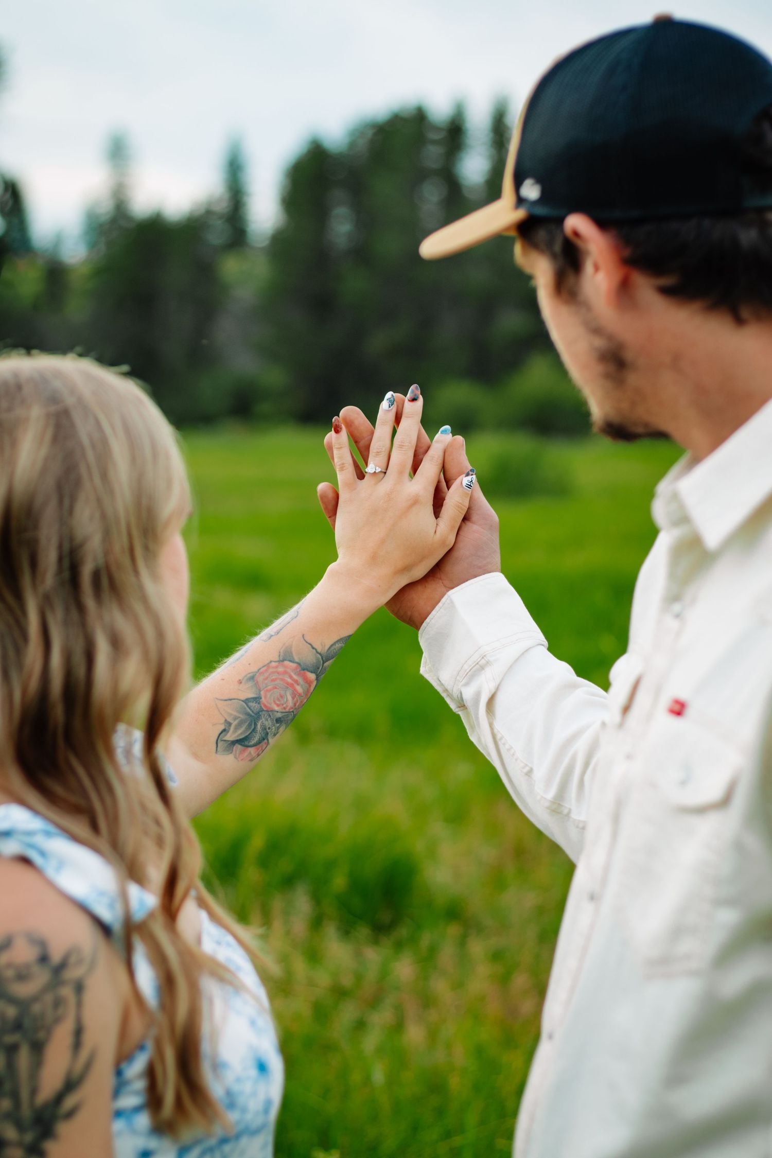 Couple holding up hands to display engagement ring in a green outdoor field setting.