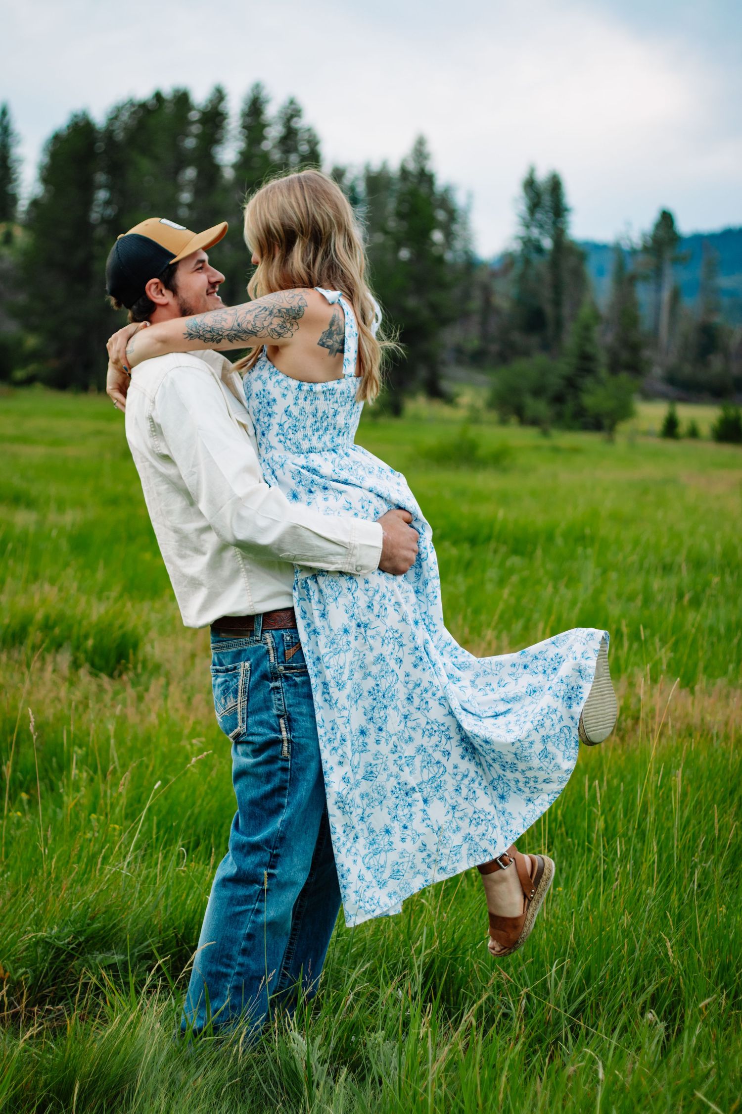 Man lifting woman in blue floral dress while embracing in a scenic green meadow with pine trees.