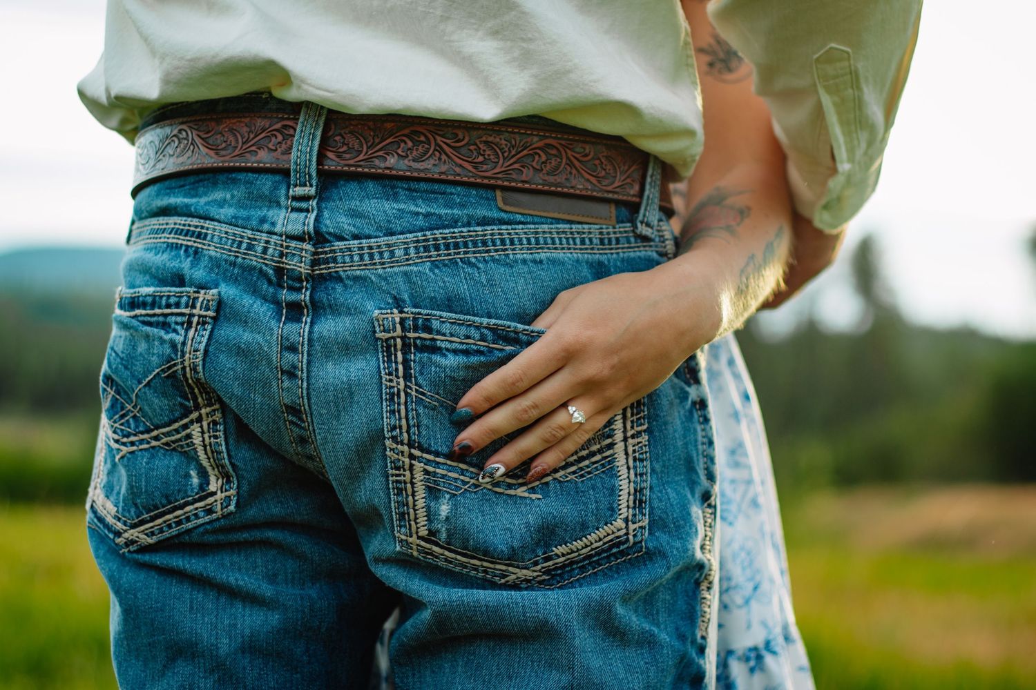 Close-up of man in jeans with decorative belt and womans tattooed hand on his back pocket.