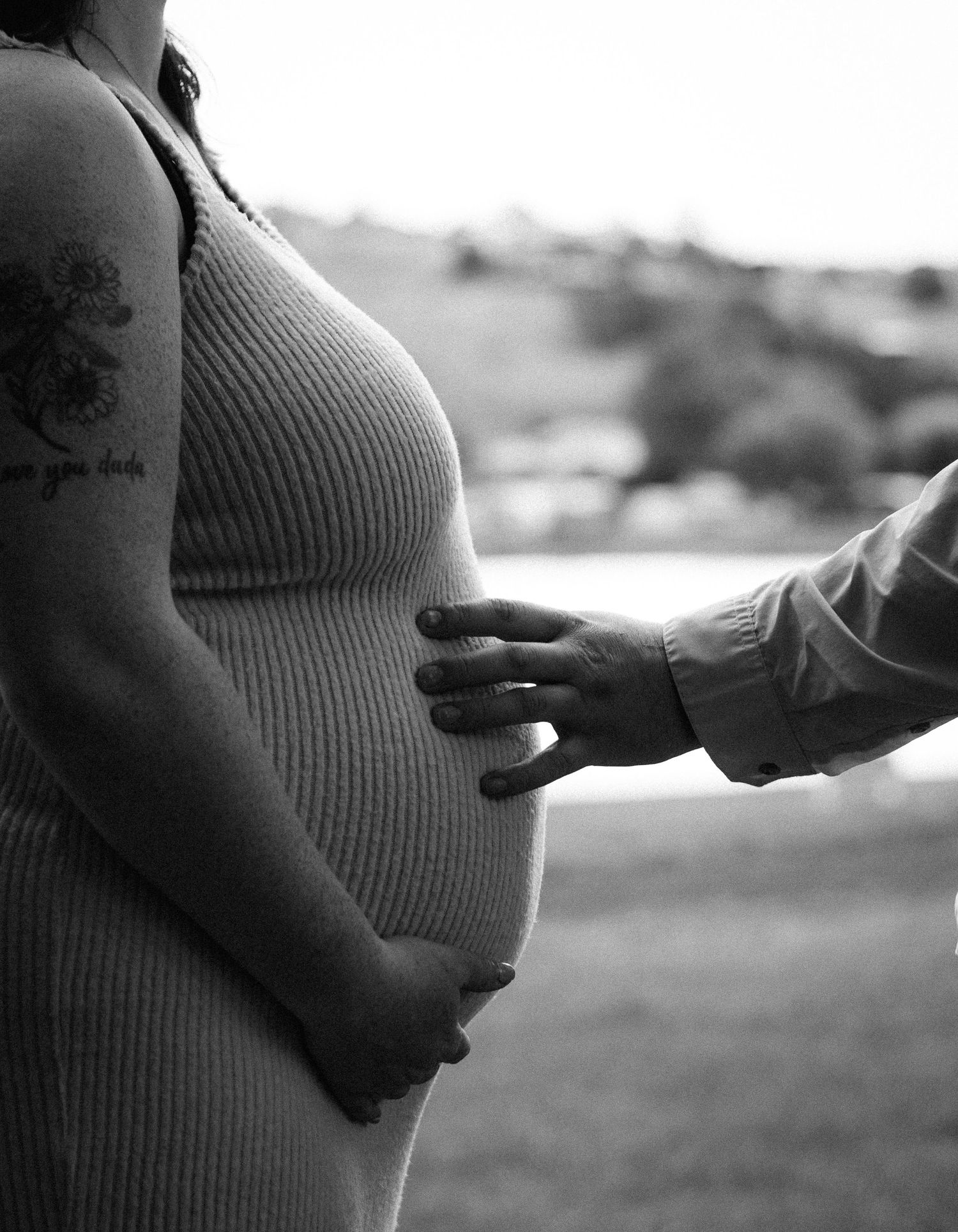 Black and white photo of pregnant woman in ribbed dress with partners hand on belly.