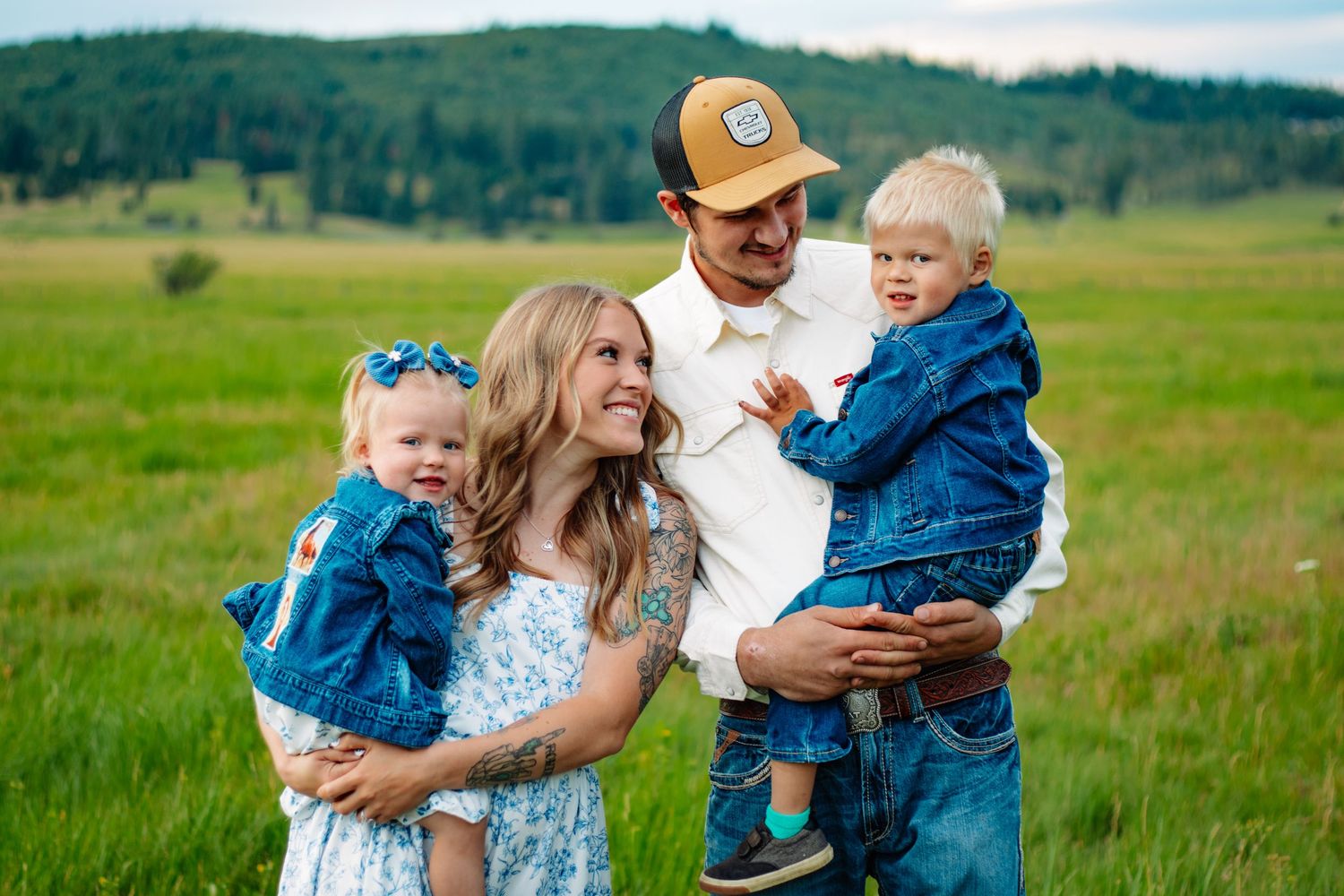 Happy family holding two toddlers in denim jackets smiling in green mountain landscape.