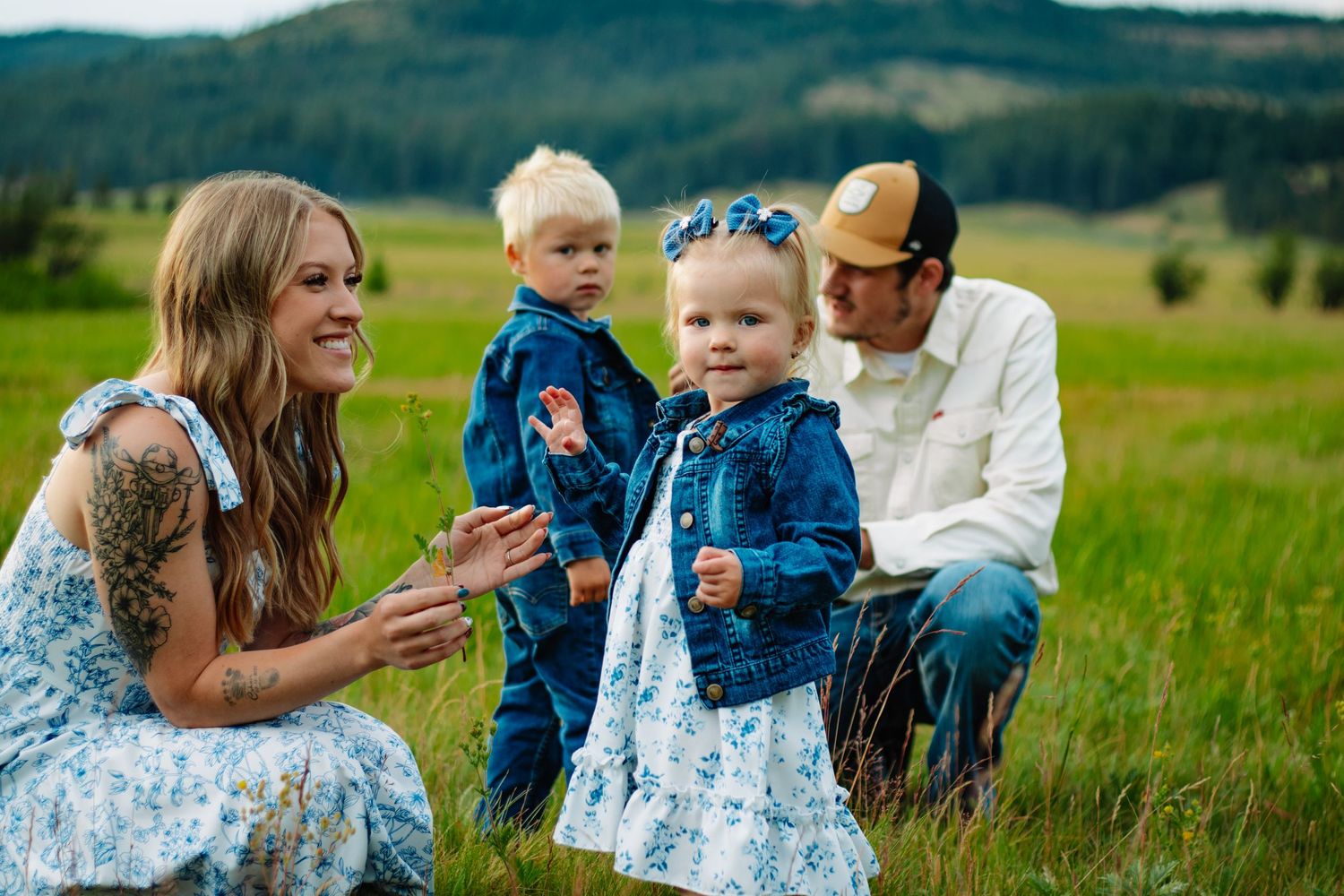 Young family with twin toddlers in matching denim outfits sitting in lush green mountain meadow.