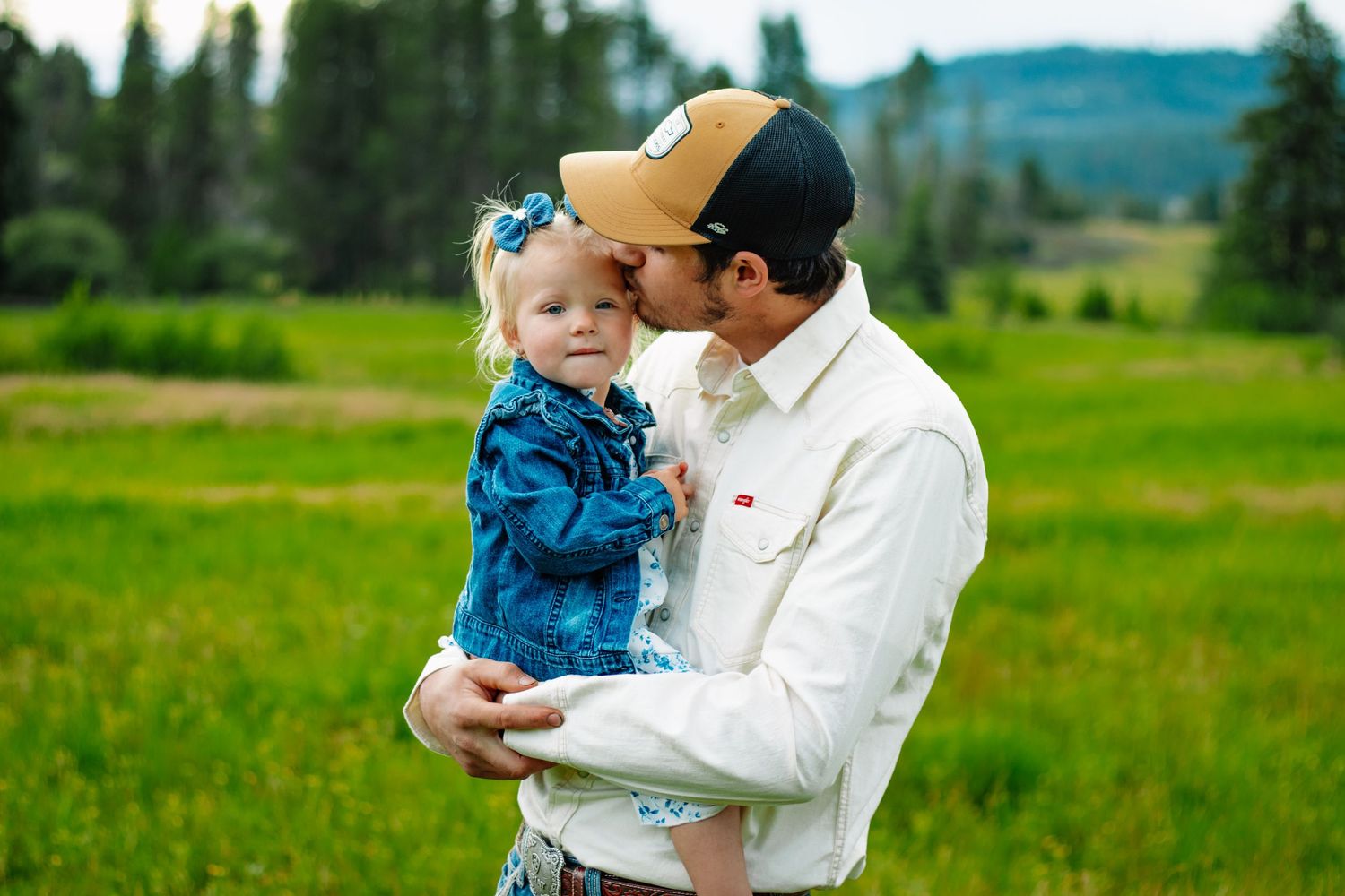Father kissing young daughter in denim jacket and blue bow in green meadow.